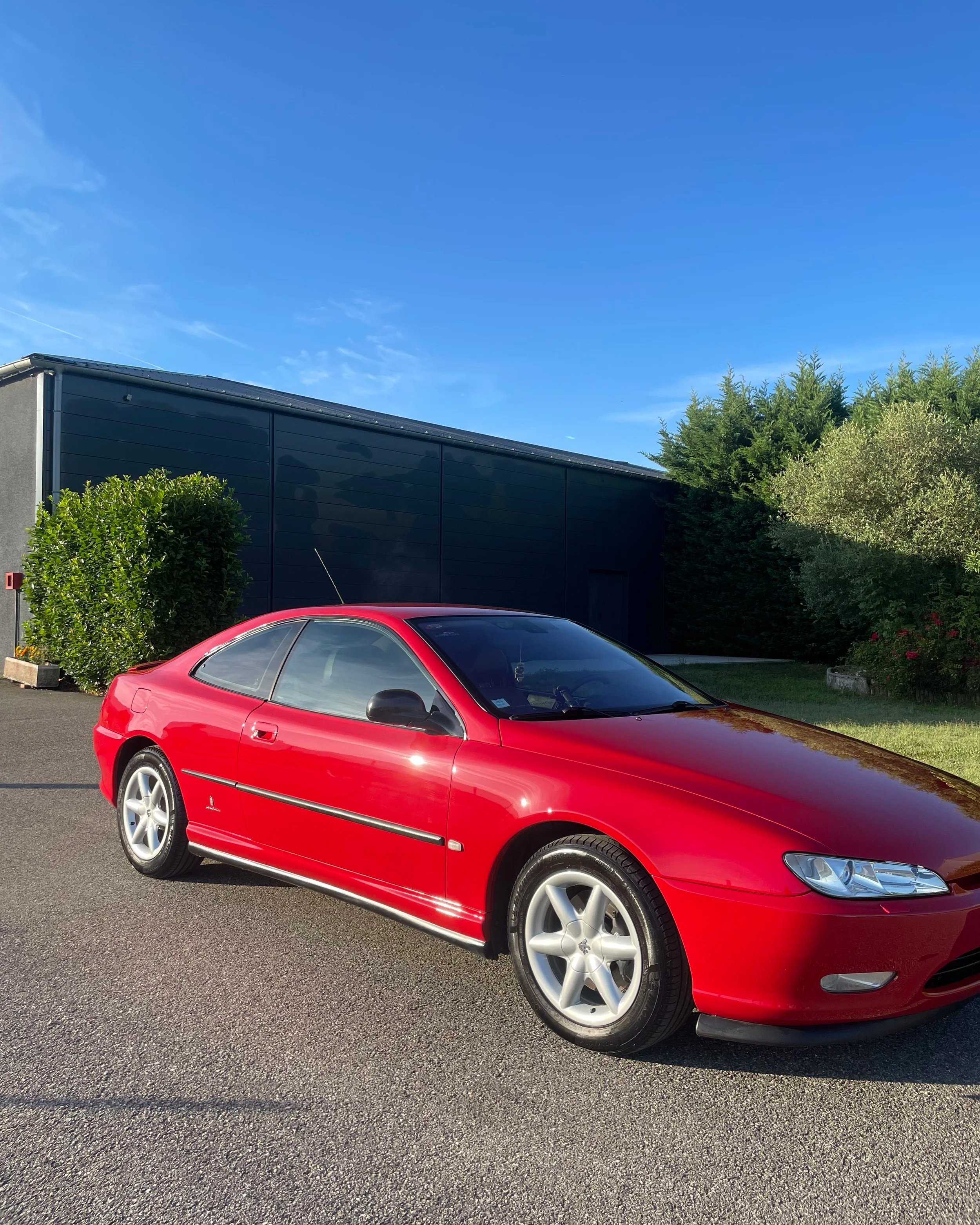 Voiture rouge stationnée sur le côté d'une route devant un bâtiment sombre et des buissons verts, avec un ciel bleu clair.
