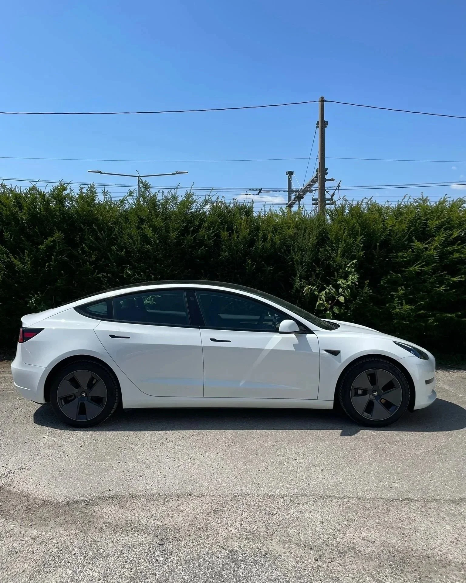 Voiture électrique blanche stationnée sur un chemin avec un fond de buissons et un ciel bleu, avec des lignes électriques en arrière-plan.