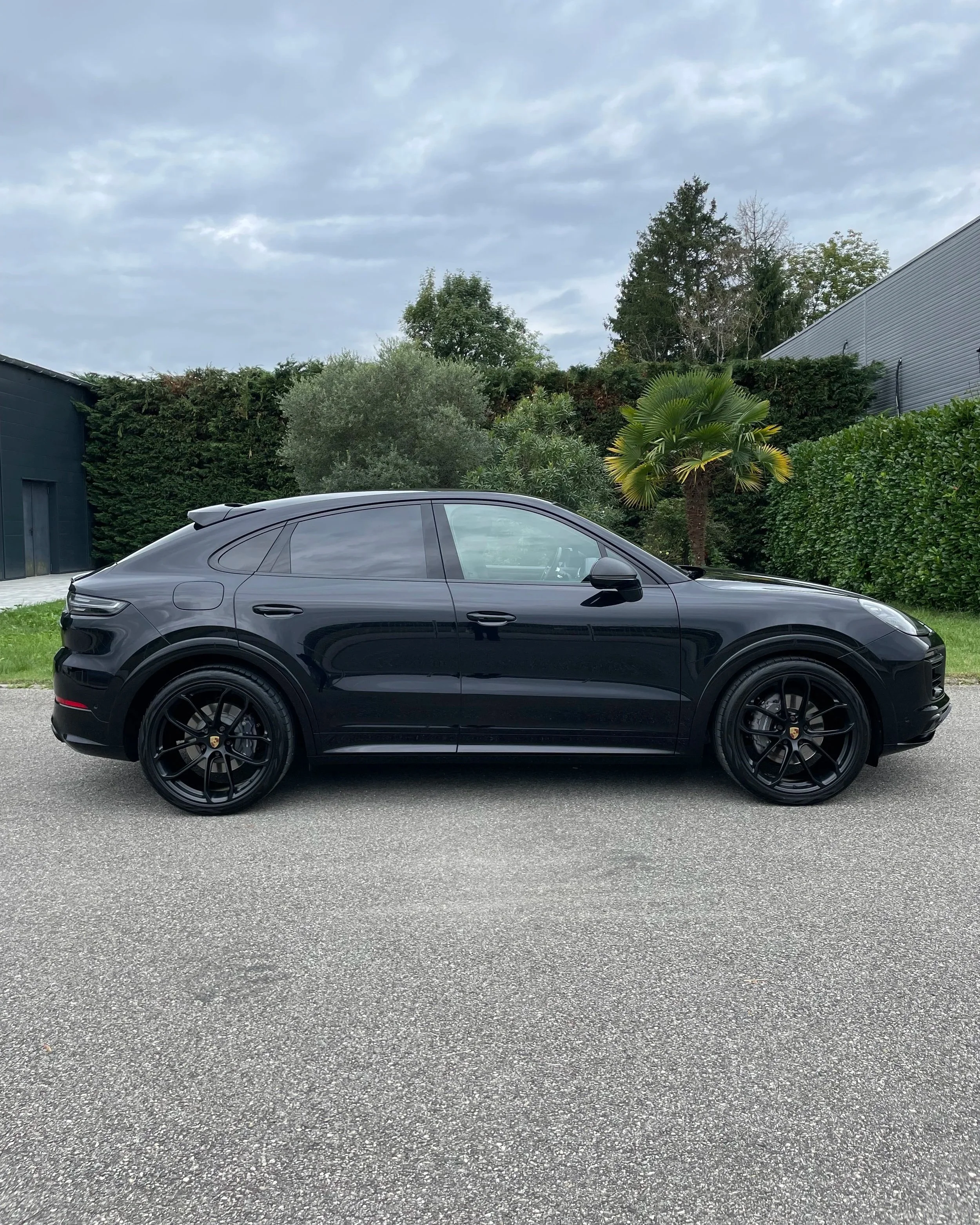 Voiture noire Porsche Cayenne coupé stationnée sur la route, avec des arbres et un ciel nuageux en arrière-plan.