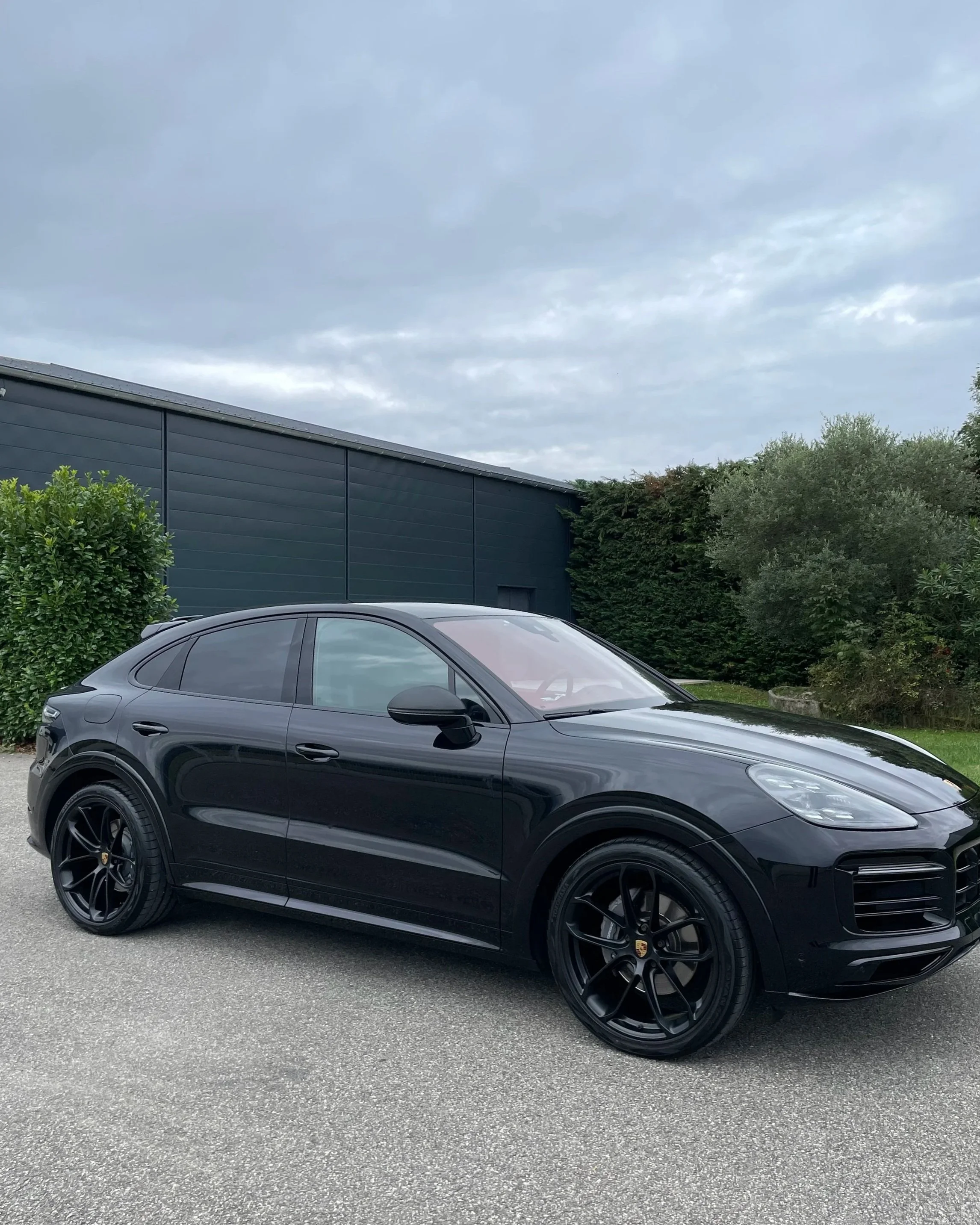 Voiture noire de luxe Porsche Cayenne sur une route, avec un bâtiment vert foncé en arrière-plan et un ciel nuageux.
