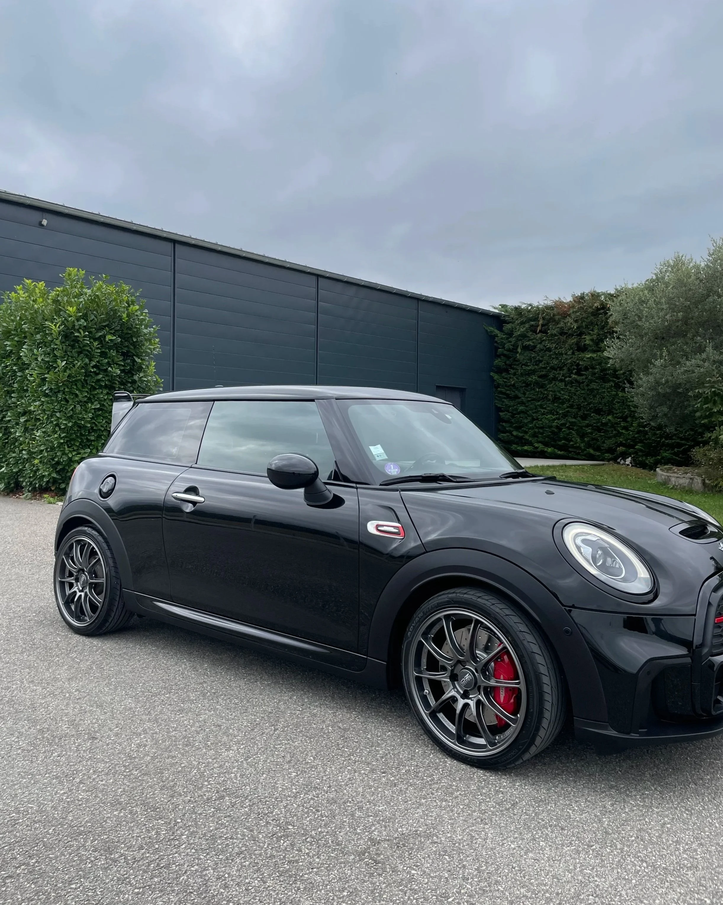 Voiture noire de modèle Mini Cooper avec jantes sportives et étriers de frein rouges, stationnée sur une surface asphaltée devant un bâtiment moderne avec des buissons et des arbres en arrière-plan, sous un ciel nuageux.