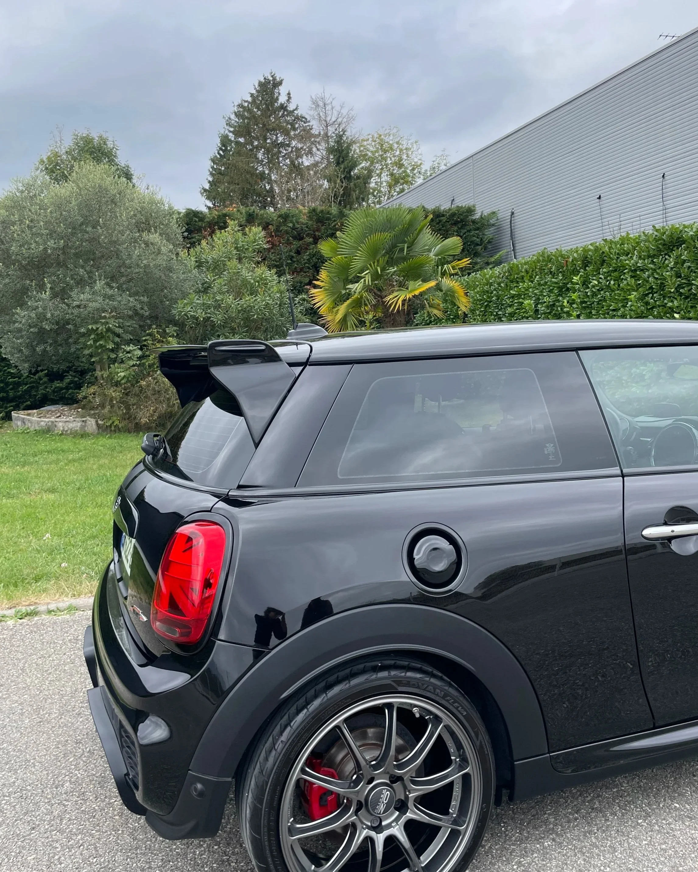 Voiture noire sport deux portes avec aileron arrière, jantes en alliage, et pneus performance, stationnée sur un trottoir à côté d'arbustes verts et d'un bâtiment industriel.
