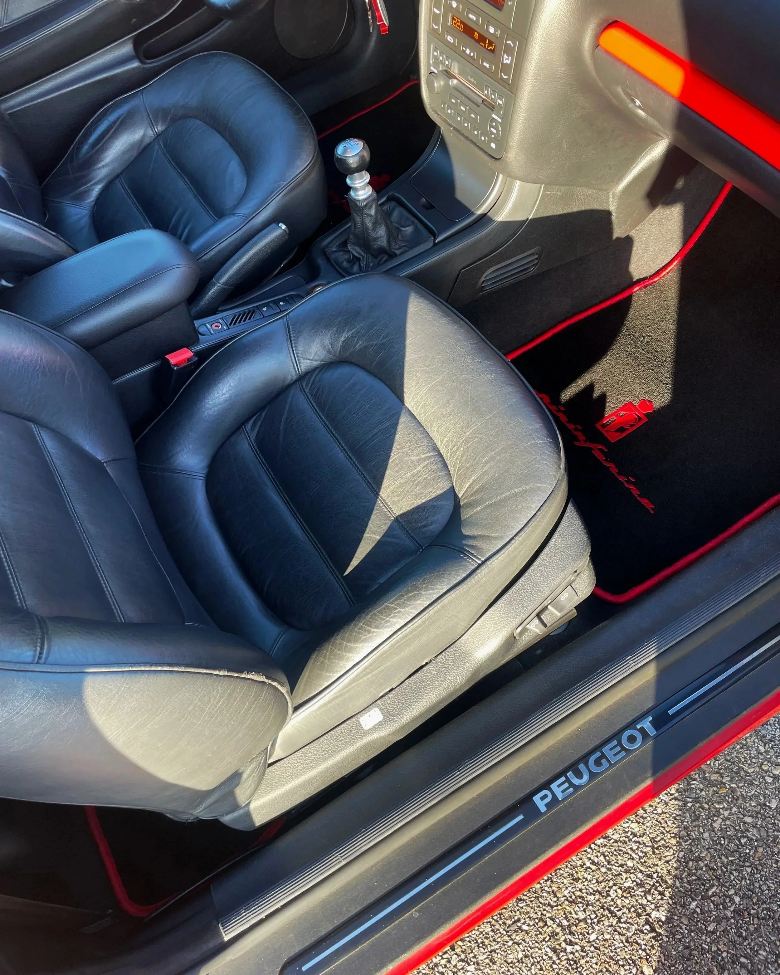 Intérieur d'une voiture Peugeot avec sièges en cuir noir, console centrale, levier de vitesse, tapis de sol noir avec le logo Peugeot en rouge et le mot "Peugeot" inscrit sur le seuil de la porte.