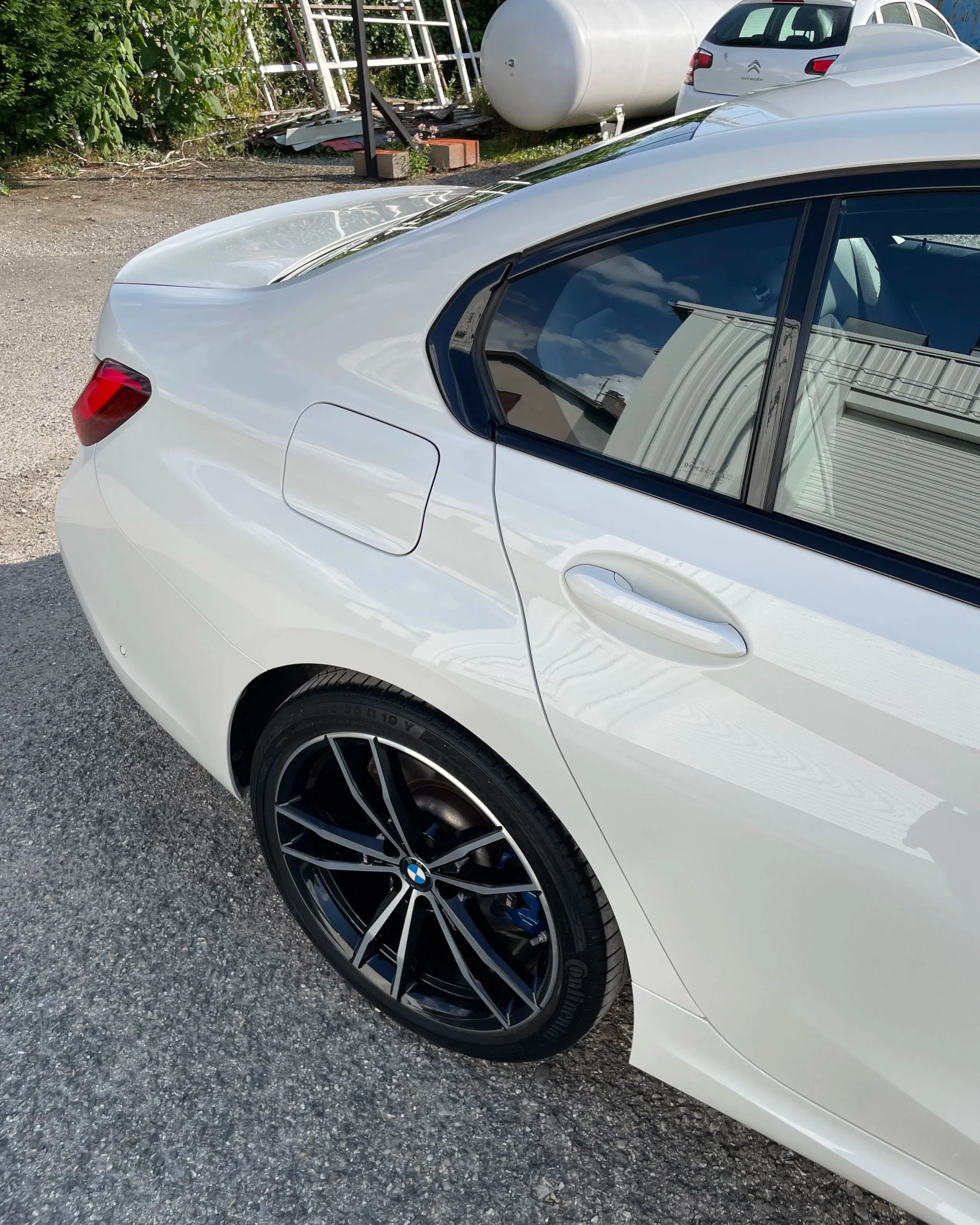 Voiture BMW blanche vue de côté, montrant la roue arrière et la porte passager, avec un fond comprenant un réservoir d'essence blanc et d'autres voitures.