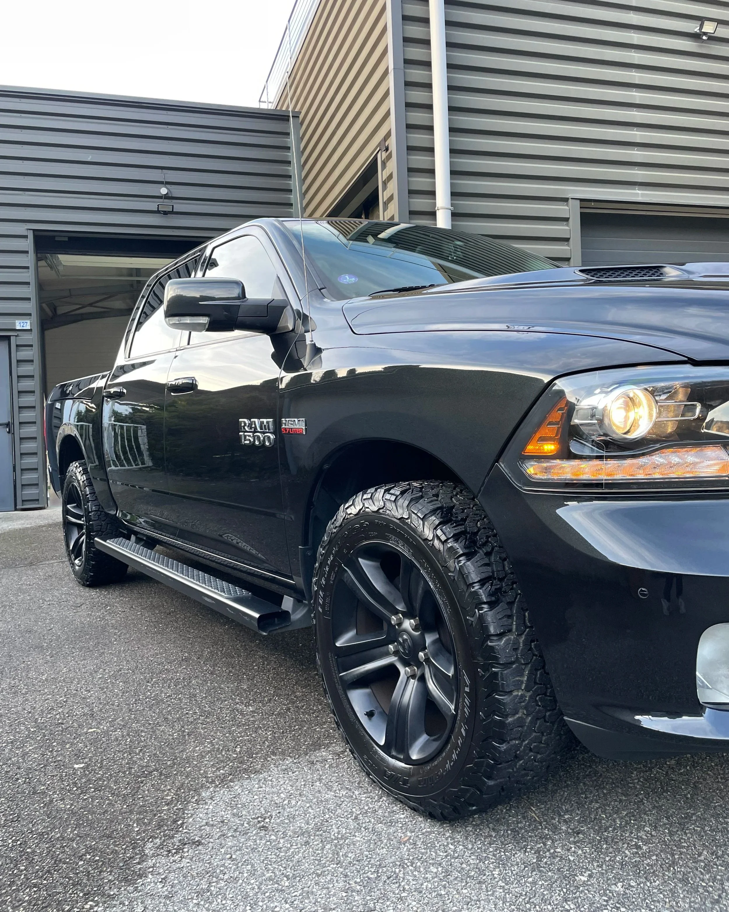 Voiture noire RAM 1500 avec roues tout-terrain, stationnée devant un bâtiment moderne en métal gris.