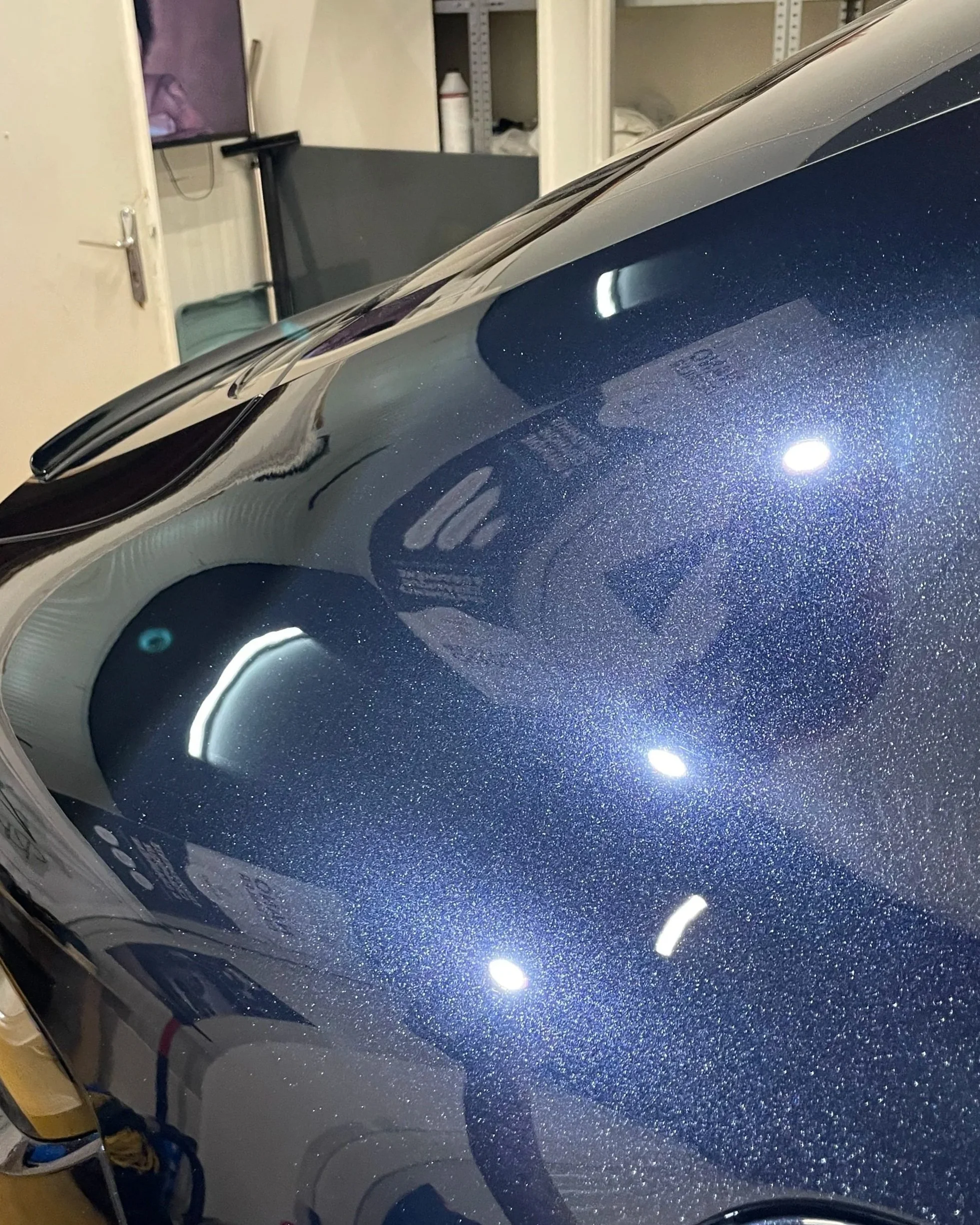 Surface de voiture en peinture métallisée bleu foncé dans un garage, avec des reflets de lumières et d'objets.