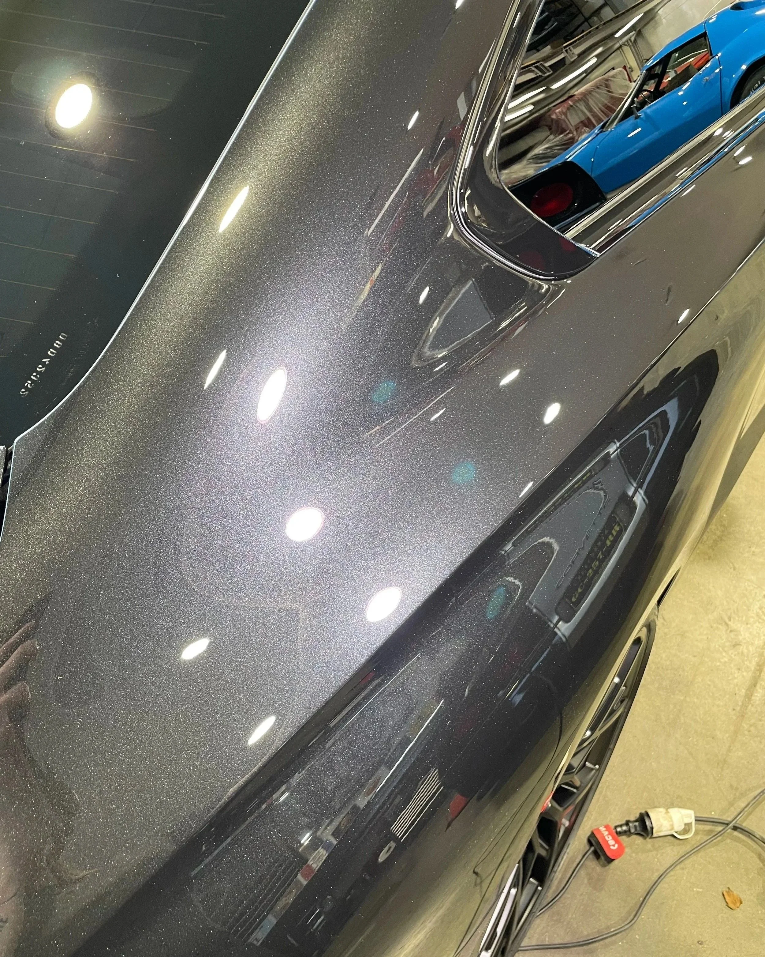 Une voiture noire avec une peinture métallisée brillante, vue de dessus dans un endroit éclairé par des lampes, avec un miroir réfléchissant le toit de la voiture et une voiture bleue en arrière-plan.