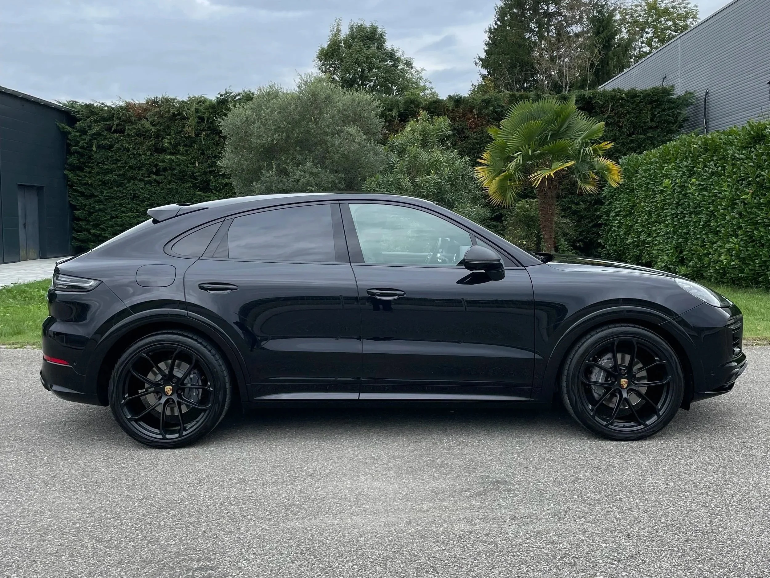 Voiture noire de luxe, modèle SUV Porsche, stationnée sur une route en extérieur, avec des arbres et une haie en arrière-plan.