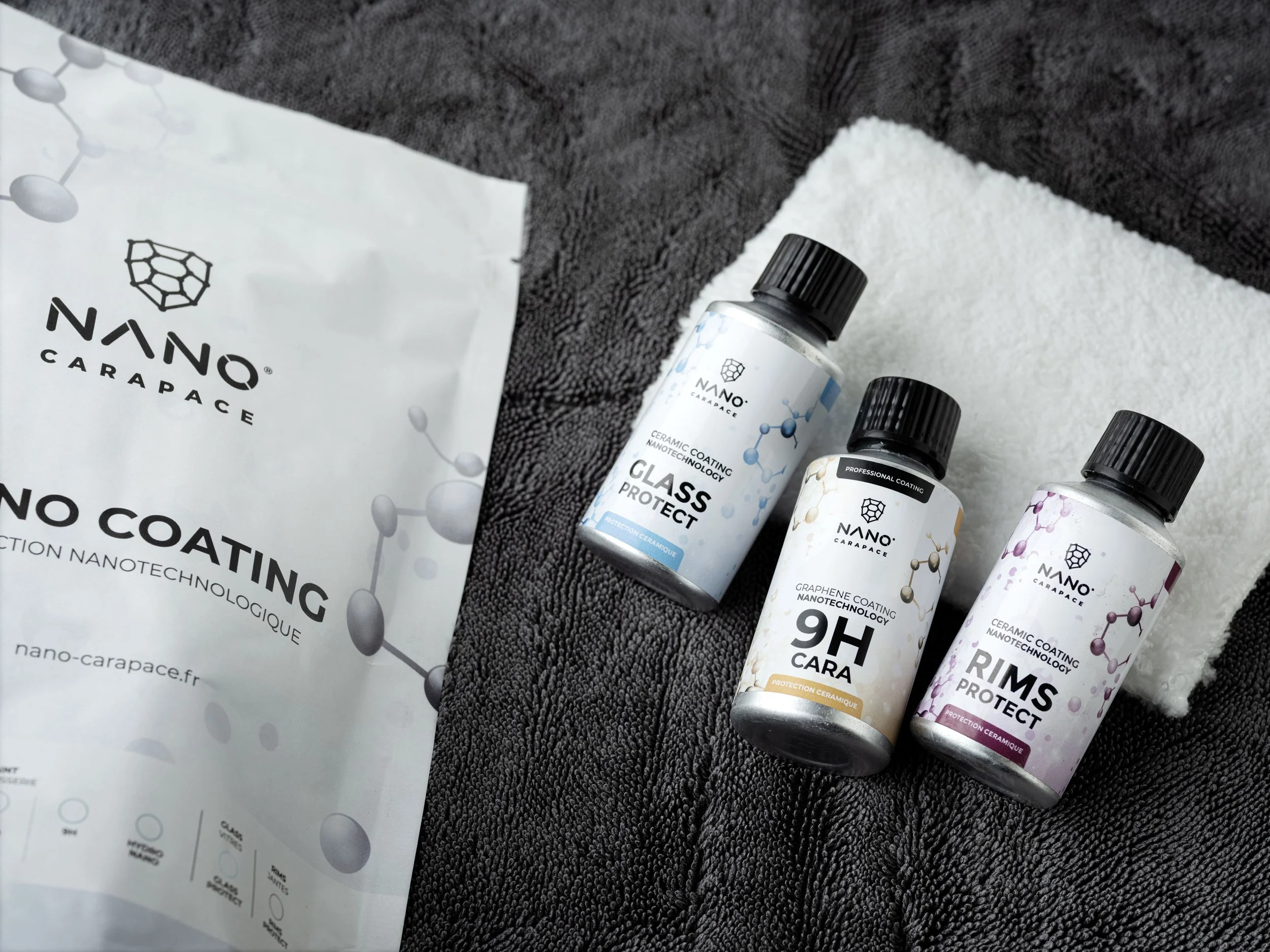 Trois flacons de protection pour le verre, le carbone et les rebords de voitures, accompagnés d'un sachet en papier de la marque Nano Carapace, tous posés sur un tapis noir