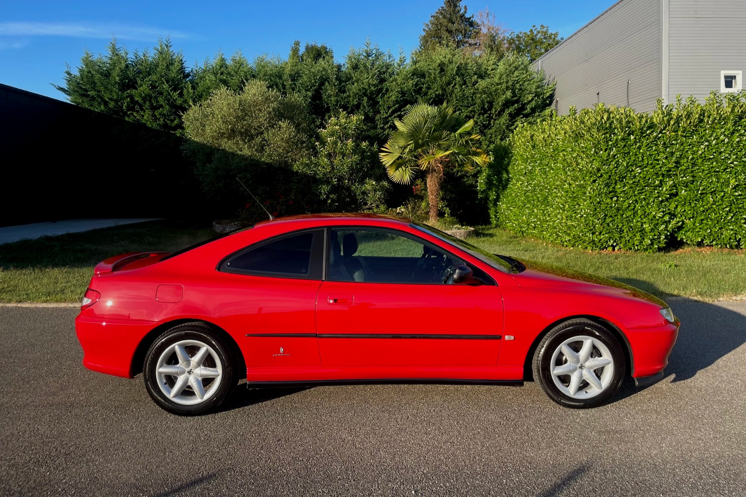 Une voiture rouge stationnée sur une route, avec un jardin verdoyant en arrière-plan et un ciel bleu clair.