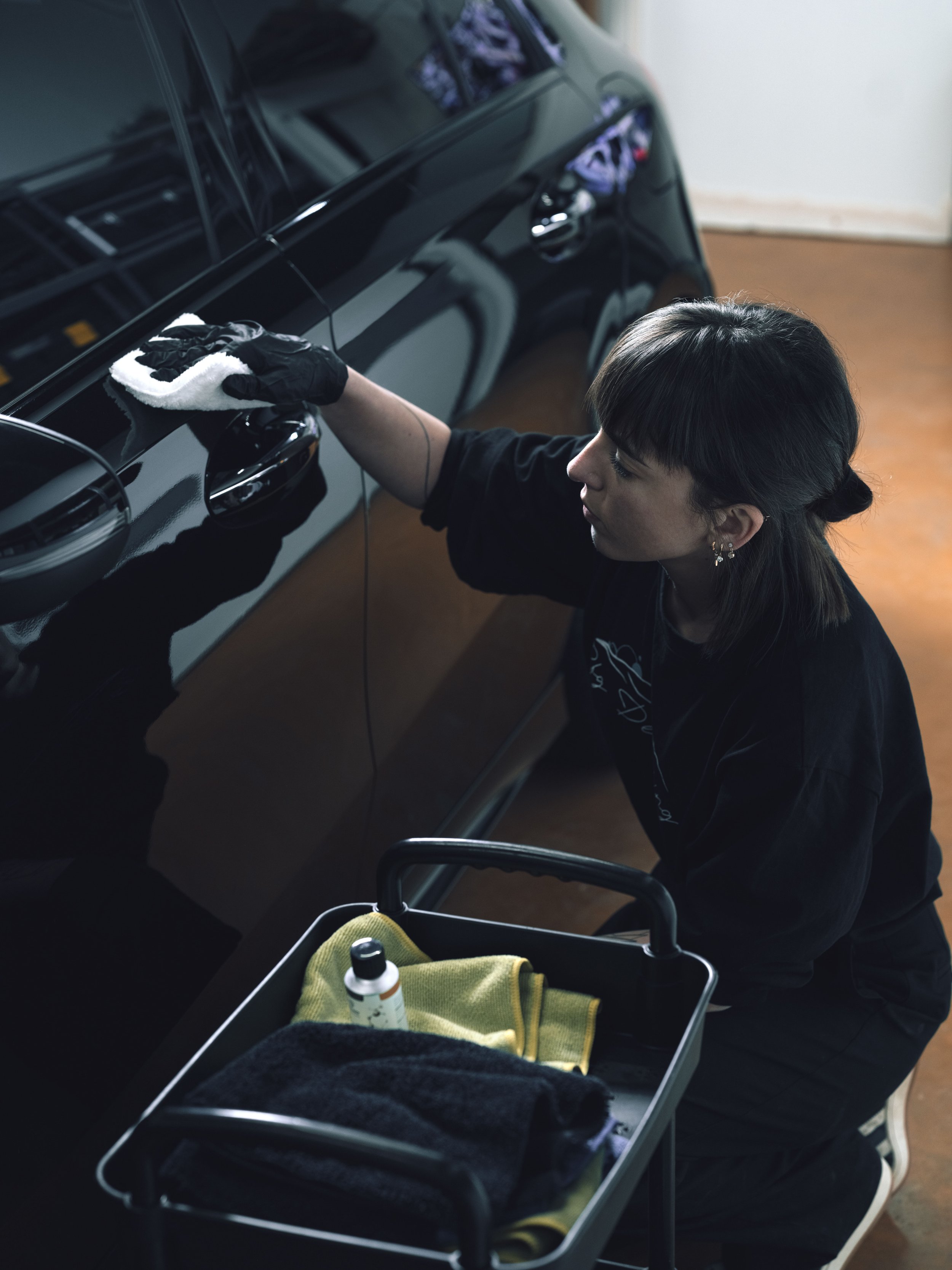 Une femme nettoie une voiture noire avec un chiffon, portant un gant noir, à l'intérieur d'un garage. Un chariot à proximité contient des serviettes, une bouteille et d'autres produits de nettoyage.