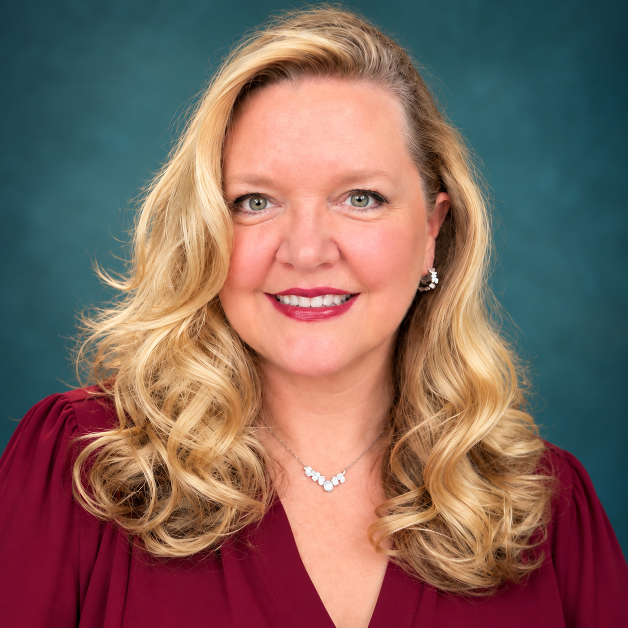 A woman with long, wavy blonde hair, wearing a maroon blouse, silver earrings, and a matching necklace, smiling against a teal background.