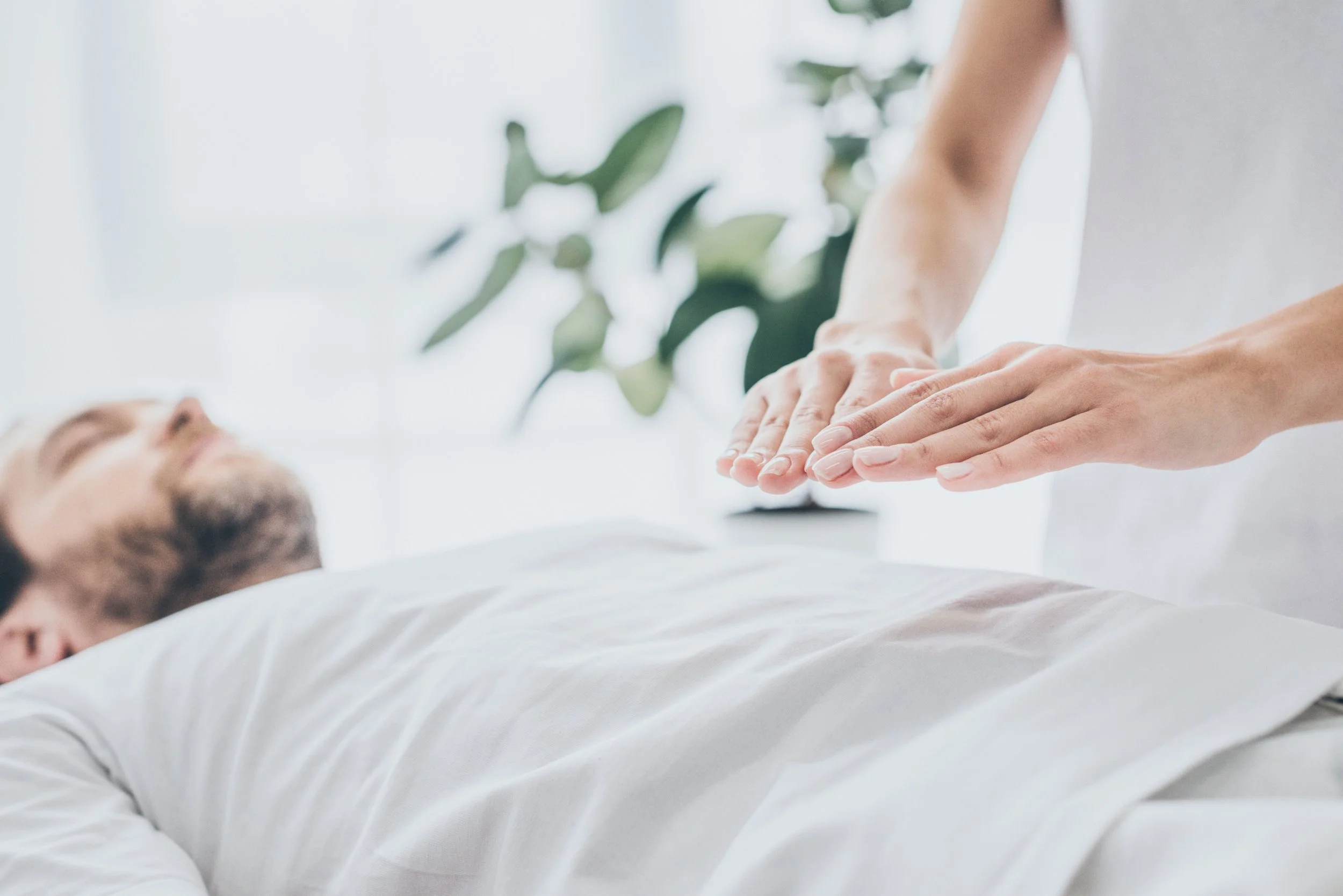 A person lying down on a bed with a white pillow, eyes closed, wearing a white shirt. A second person is performing chest compressions on them during a medical emergency. The background is bright with white and green elements, including a plant.
