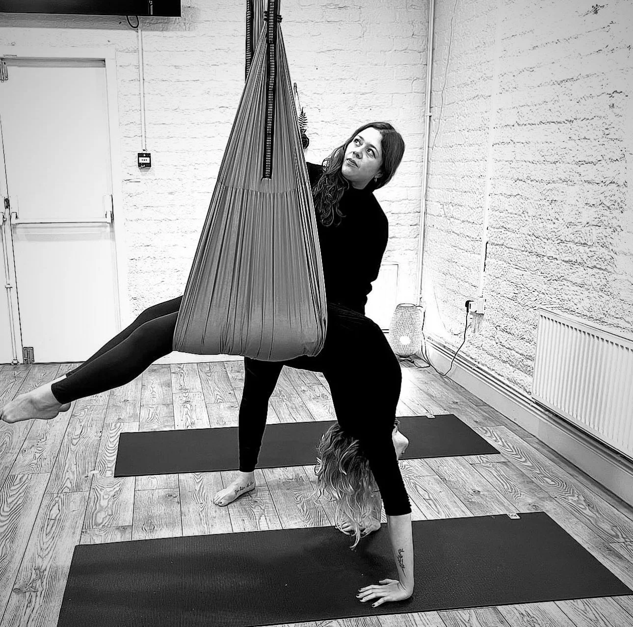 Two women practicing aerial yoga in a studio, with one woman upside down in a handstand and the other woman using aerial silk to support her legs.