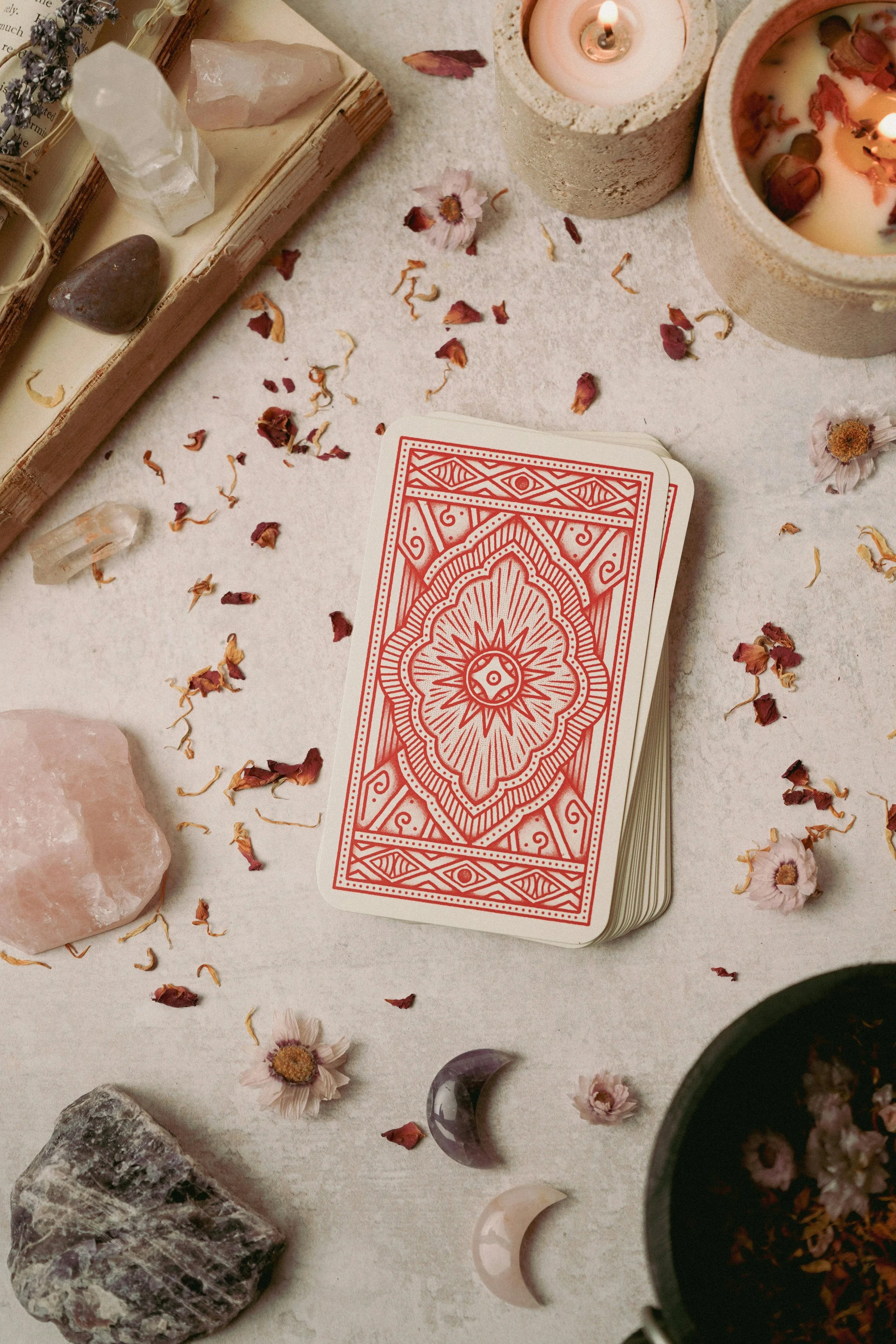 A deck of tarot cards with a red back design on a white surface surrounded by candles, crystals, dried flowers, and crescent moon ornaments.