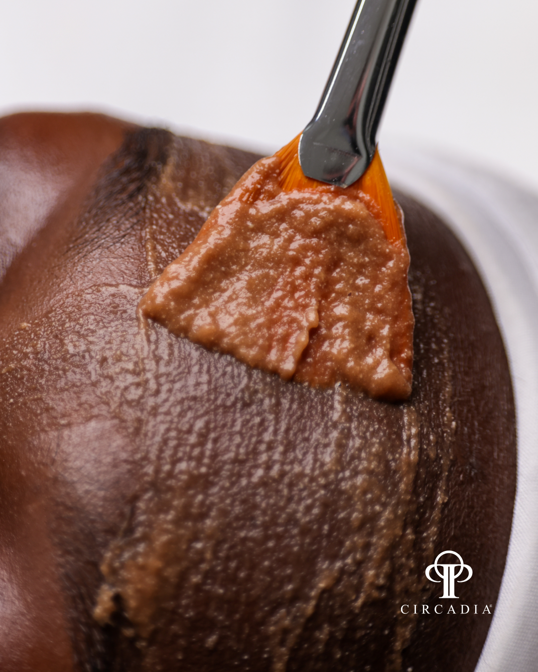 A clay facial mask being applied to a African American women cheek with a masking brush. Circadia Cocoa Enzyme Facial.  The mask is thick and brown in color, covering the skin.