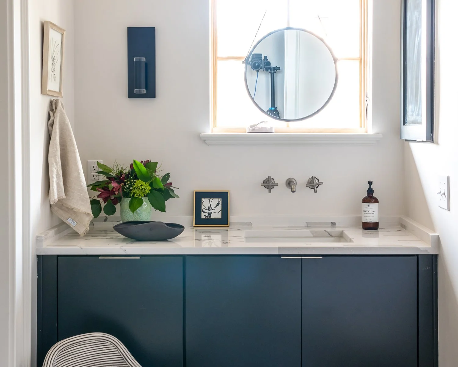 Bathroom vanity with a marble countertop, dark blue cabinets, a vase of flowers, a framed picture, a sink, a wall-mounted faucet, a bottle of soap, and a round mirror above the window.