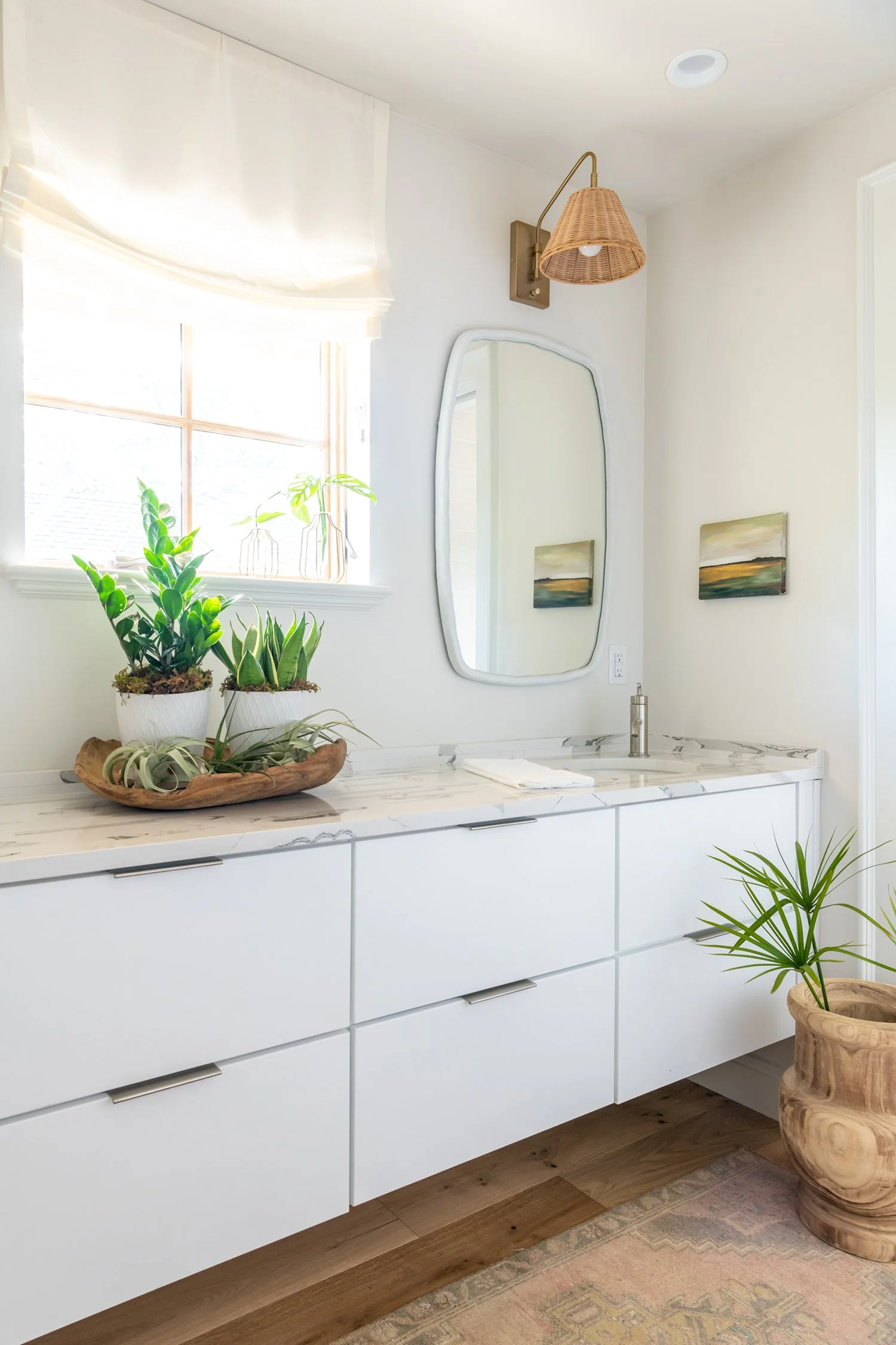 Bathroom vanity with white cabinets, marble countertop, a mirror, potted plants, and a window with a white rolled-up shade