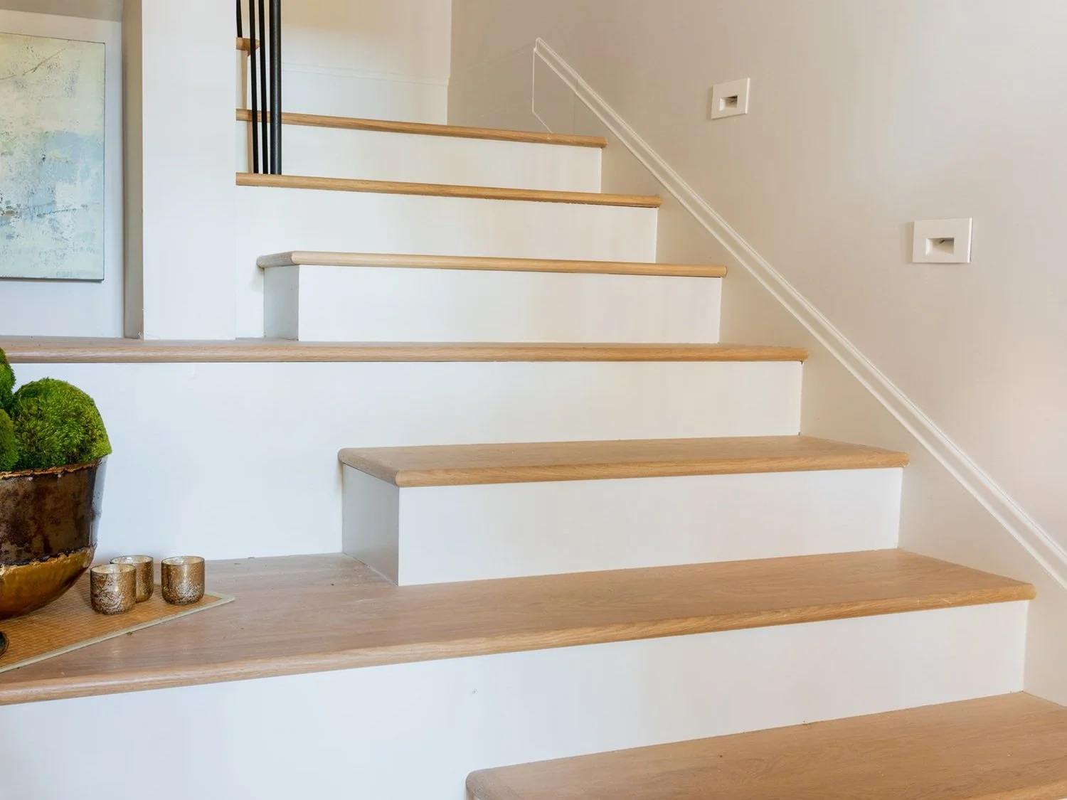 Wooden staircase with a potted plant and decorative candles at the bottom, white walls, and framed artwork on the left.
