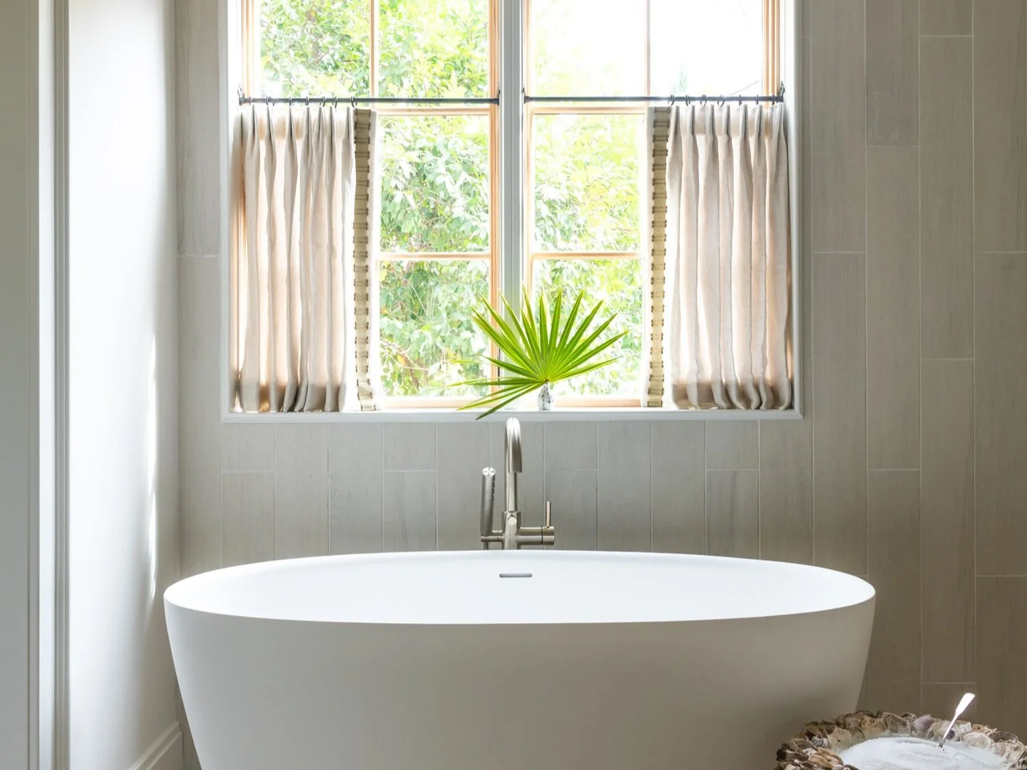 A modern bathroom with a white freestanding bathtub positioned in front of a large window with beige curtains. A green palm leaf decorates the windowsill, and sunlight filters through the window, illuminating the room.