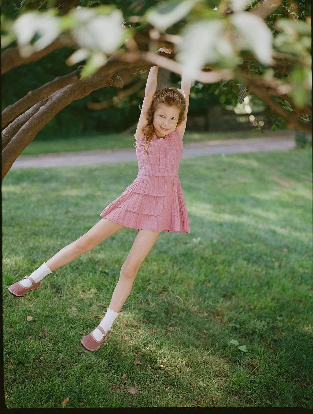 A young girl hanging from a tree branch in a grassy park, wearing a pink dress and brown shoes with white socks.