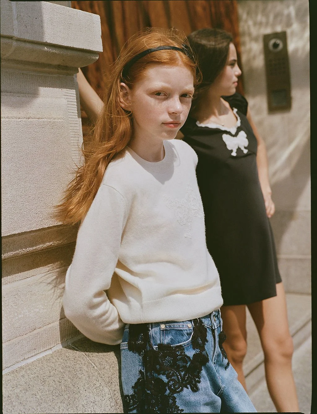 A young girl with red hair and a black headband leaning against a stone wall, wearing a white sweater and denim shorts with black lace details. In the background, a woman with dark hair in a black dress stands next to her, on a city sidewalk.