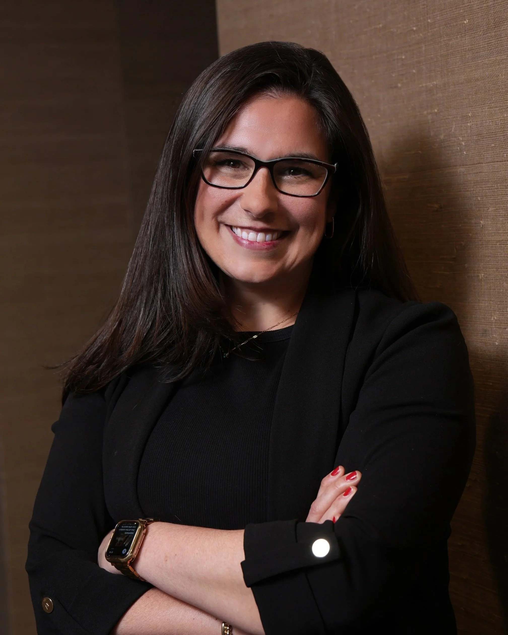 Smiling woman with dark hair, wearing hoop earrings, in front of gray background.