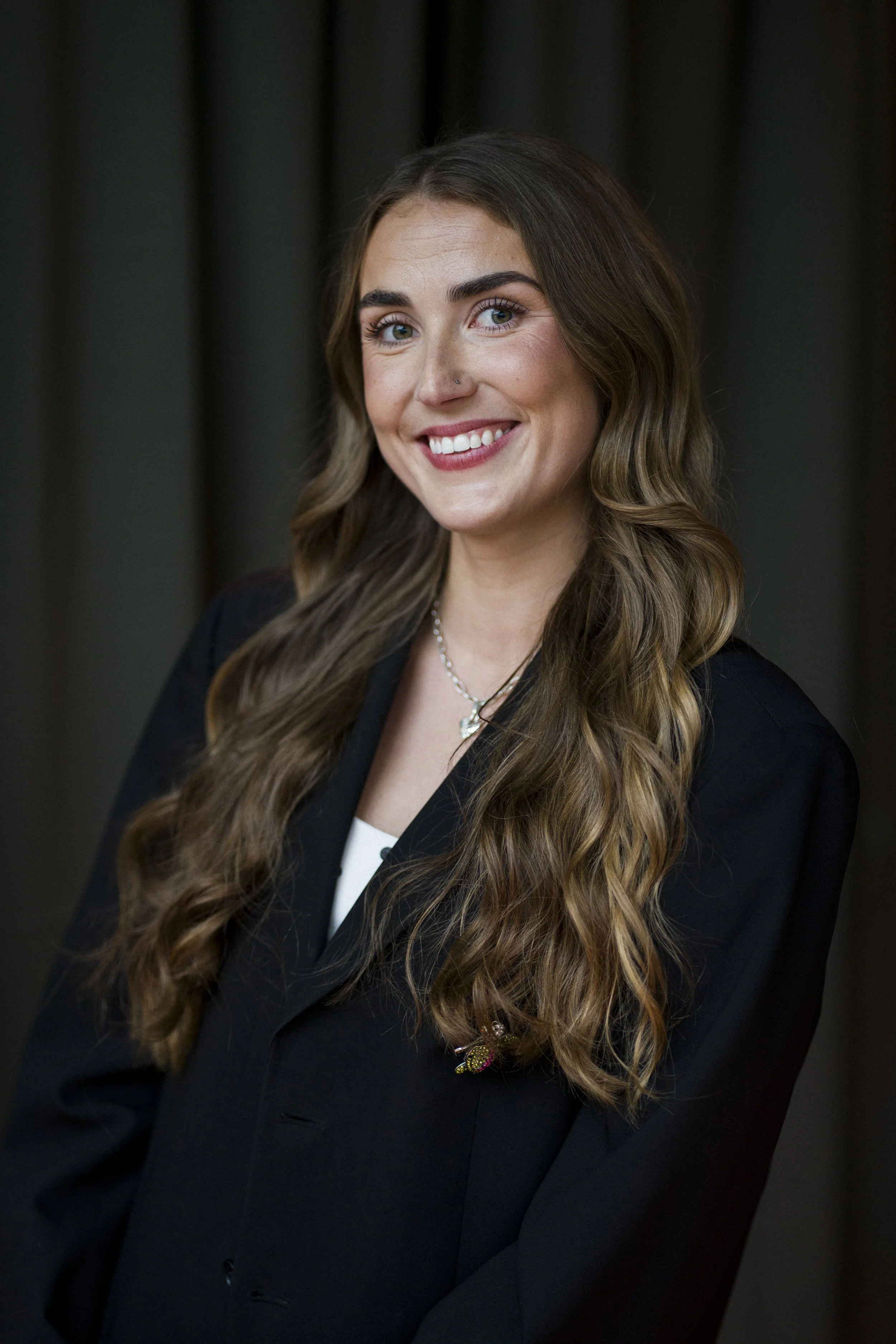 Freja Strecker partner i Pift. Profilbillede af en smilende ung kvinde med langt, krøllet brunt hår, iført sort blazer og en skjorte, står mod en mørk baggrund.