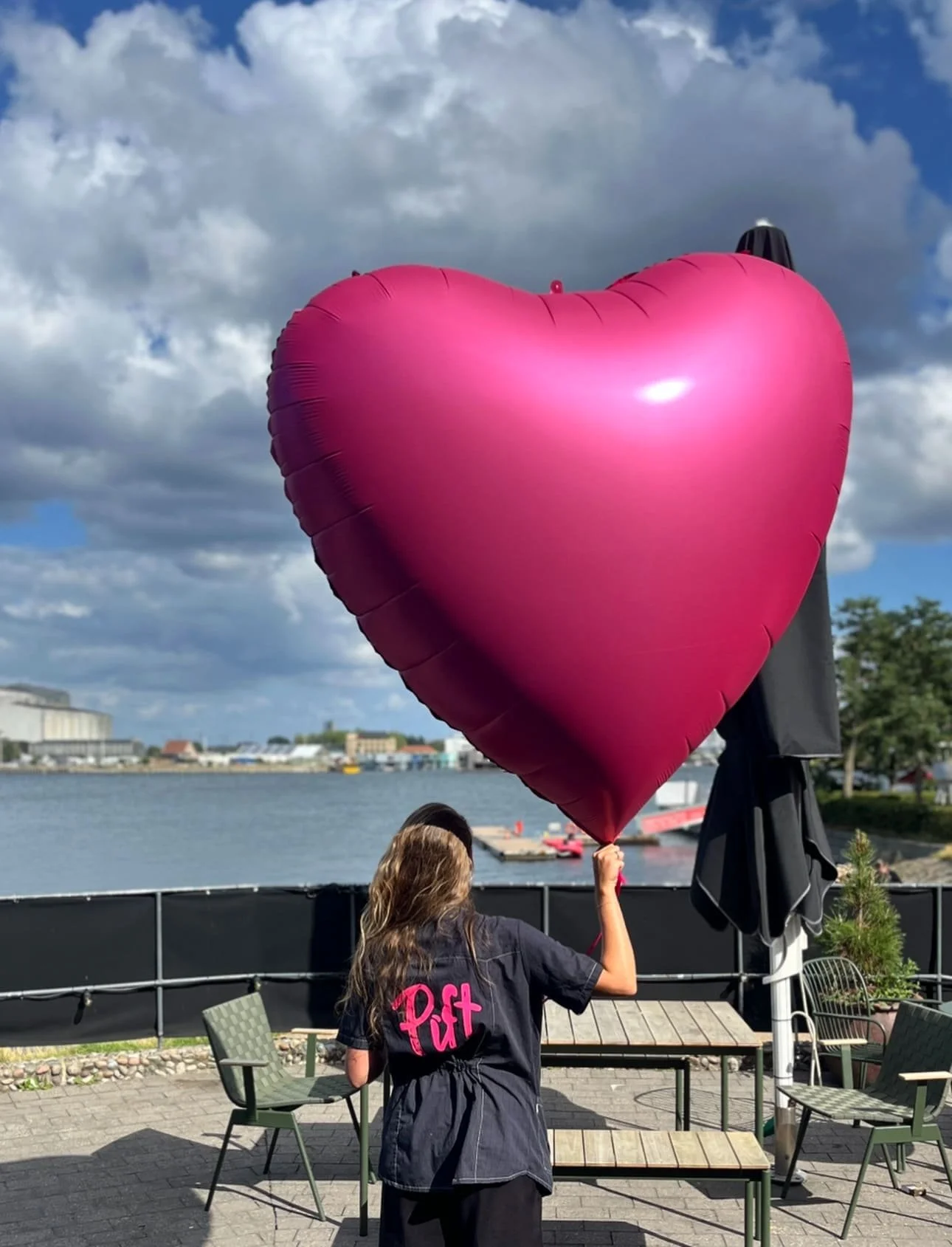 En person holder en stor, pink, hjerteformet ballon ud mod en vandkrop med en by i baggrunden og en skyet himmel.
