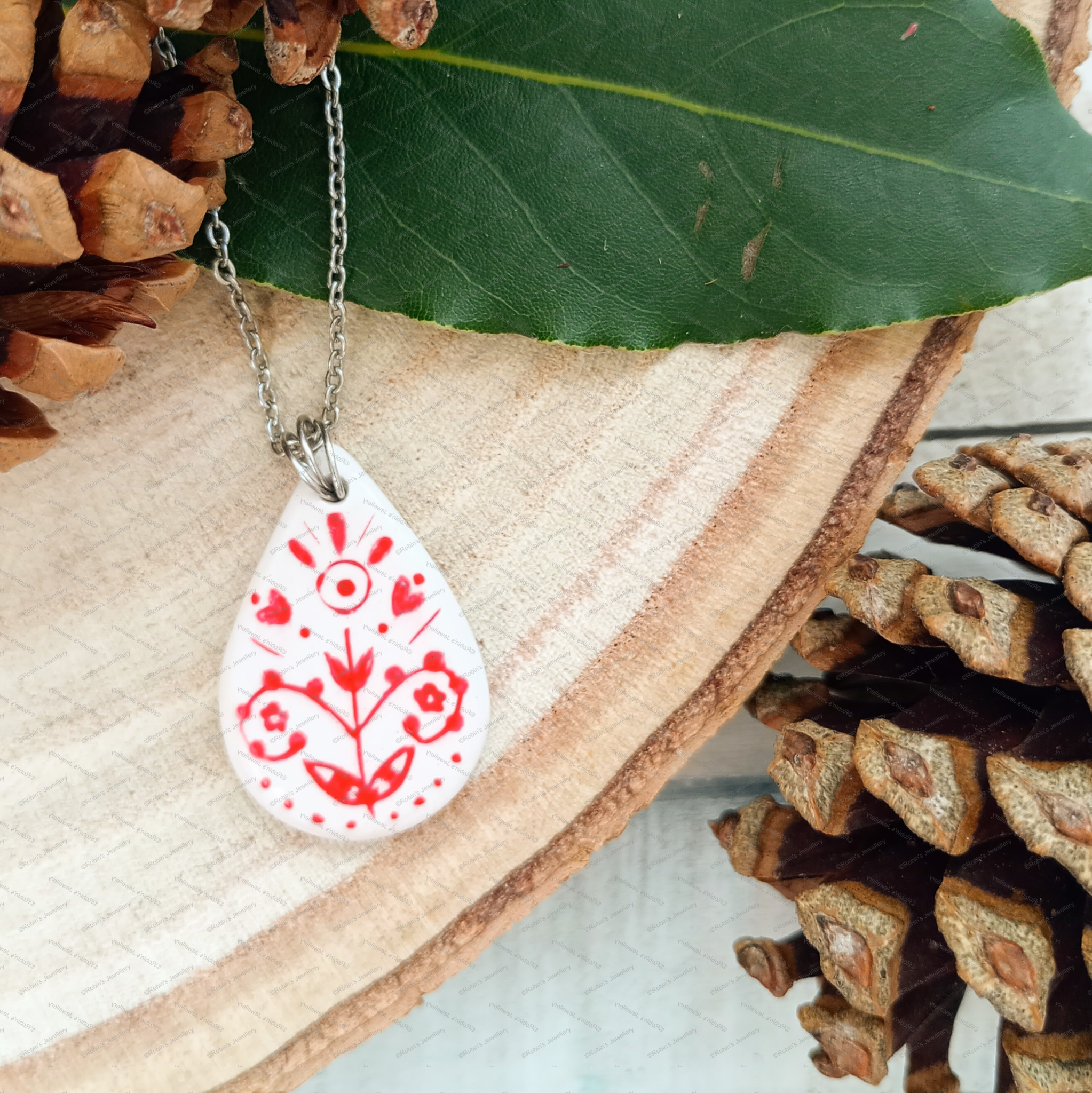 A teardrop-shaped white pendant with red floral and line art design hanging on a silver coloured chain.