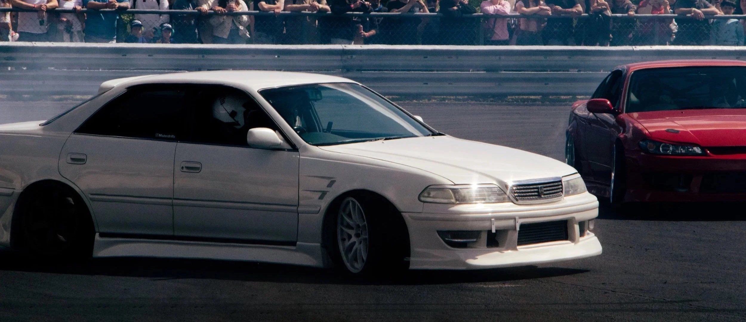 A white modified sports car racing on a track with spectators watching behind a fence, accompanied by a red car.