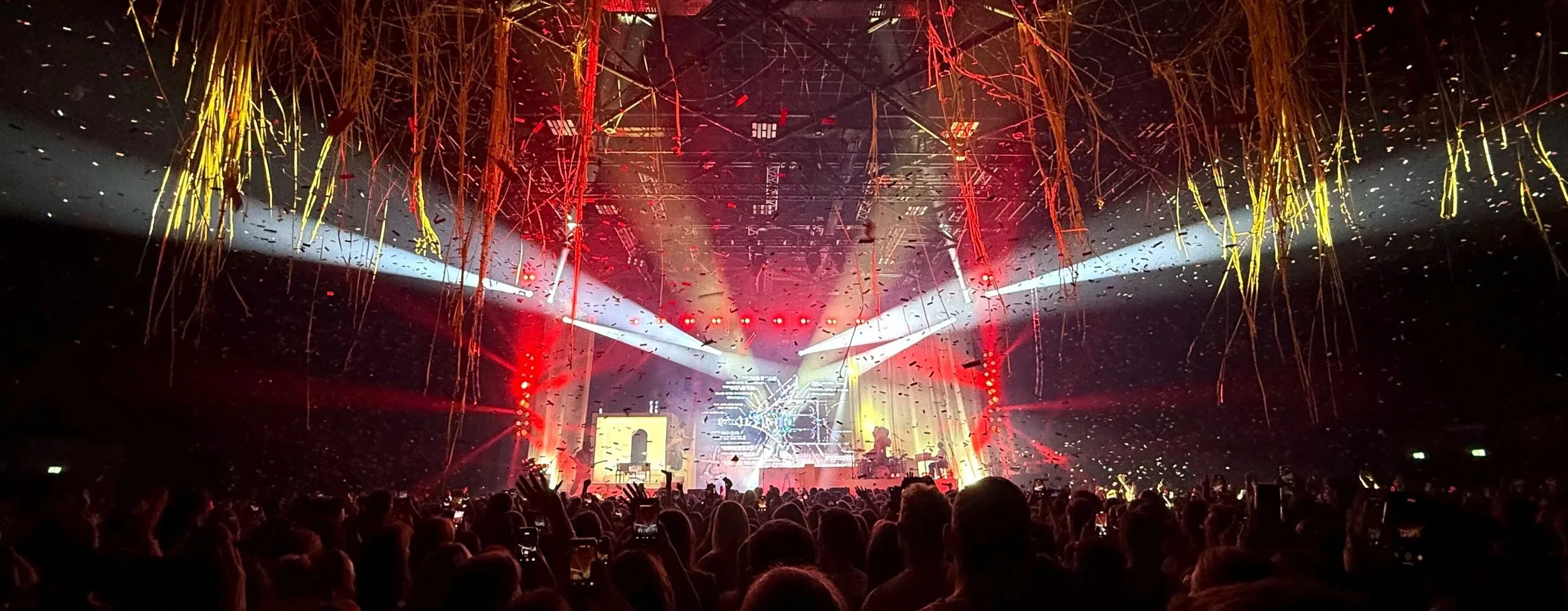 Crowd at a concert with colorful stage lights, beams, confetti, and streamers hanging from the ceiling.