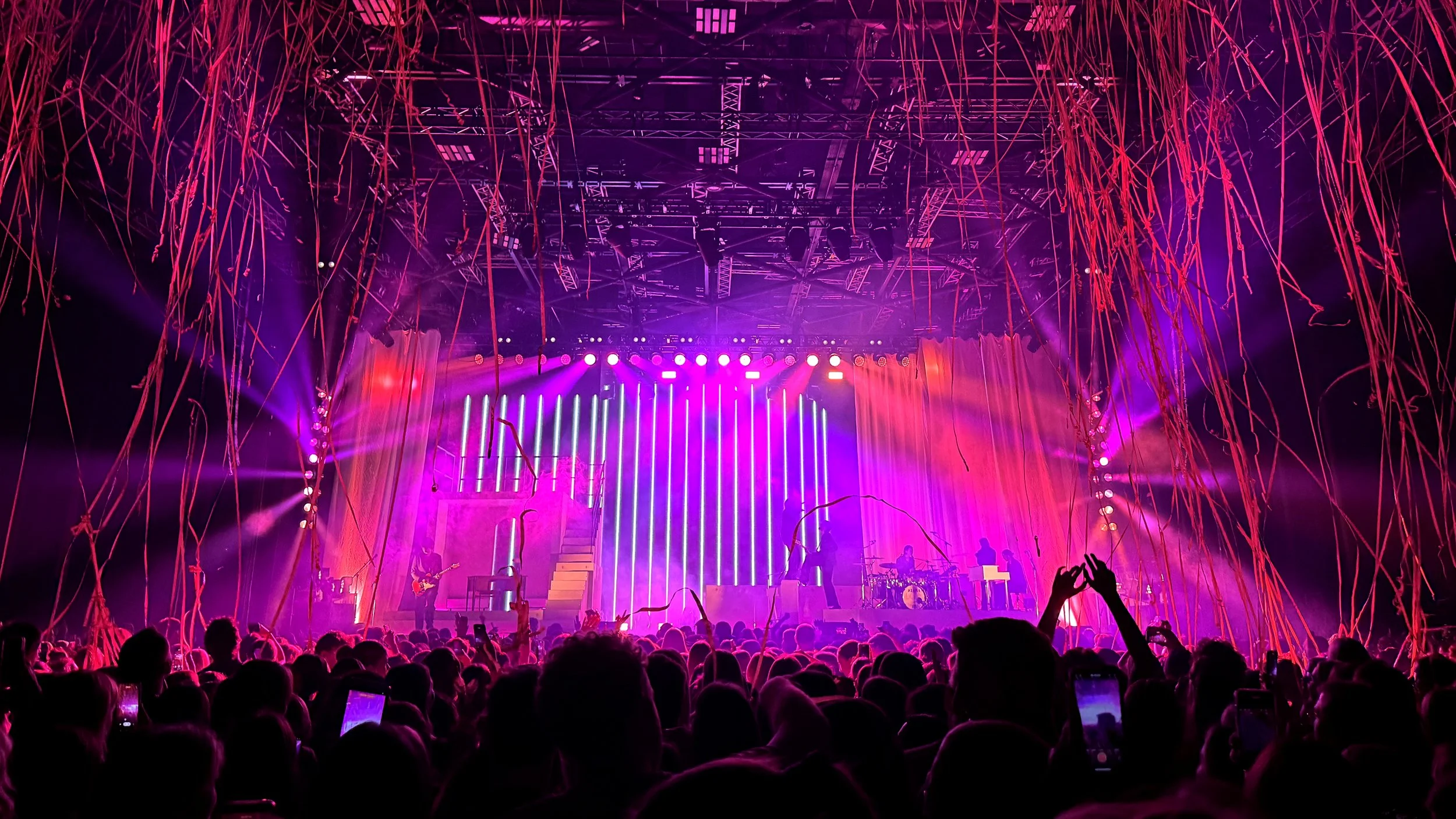 A live concert scene with a stage illuminated by vibrant pink and purple lights, surrounded by hanging red streamers, with silhouettes of musicians performing and a crowd raising hands, capturing an energetic atmosphere.