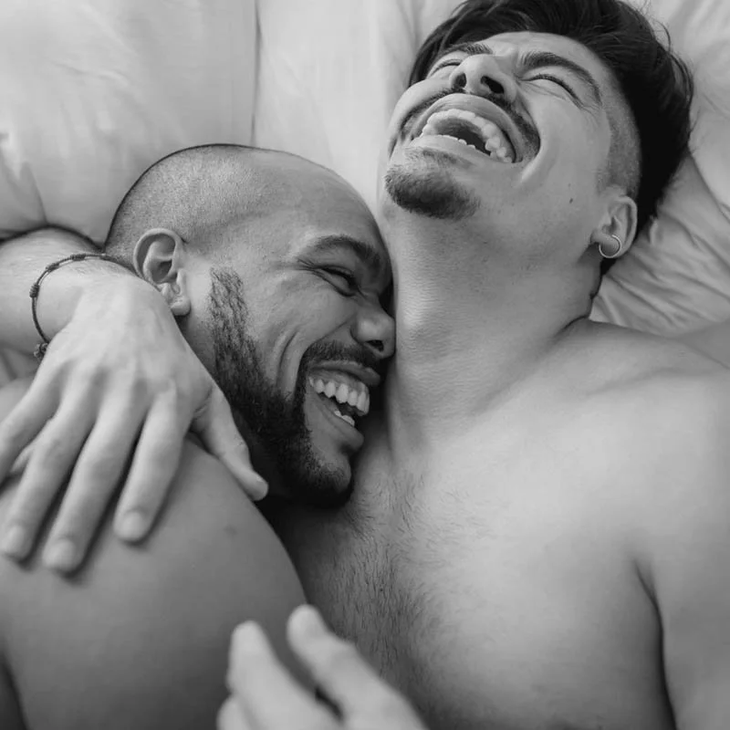 Two men are lying on a bed, embracing and smiling happily. One man has a shaved head and a beard, and the other has dark hair and a mustache.