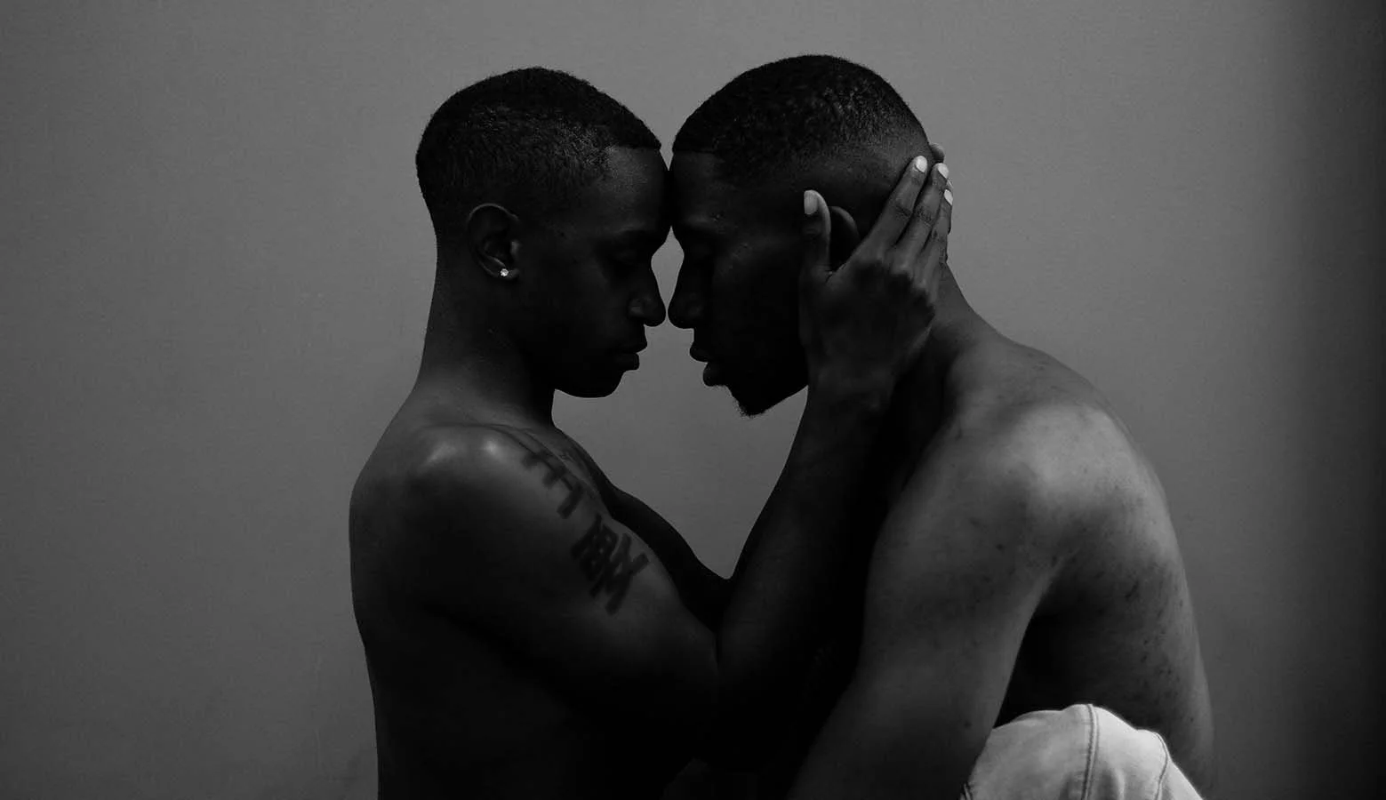 Two shirtless men, one with tattoos and earrings and the other with a trimmed beard, lean their foreheads together and hold each other's faces gently in an intimate pose in black and white.