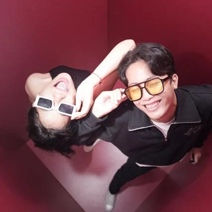 Two young people smiling and wearing sunglasses, in a red corner, taking a playful selfie.