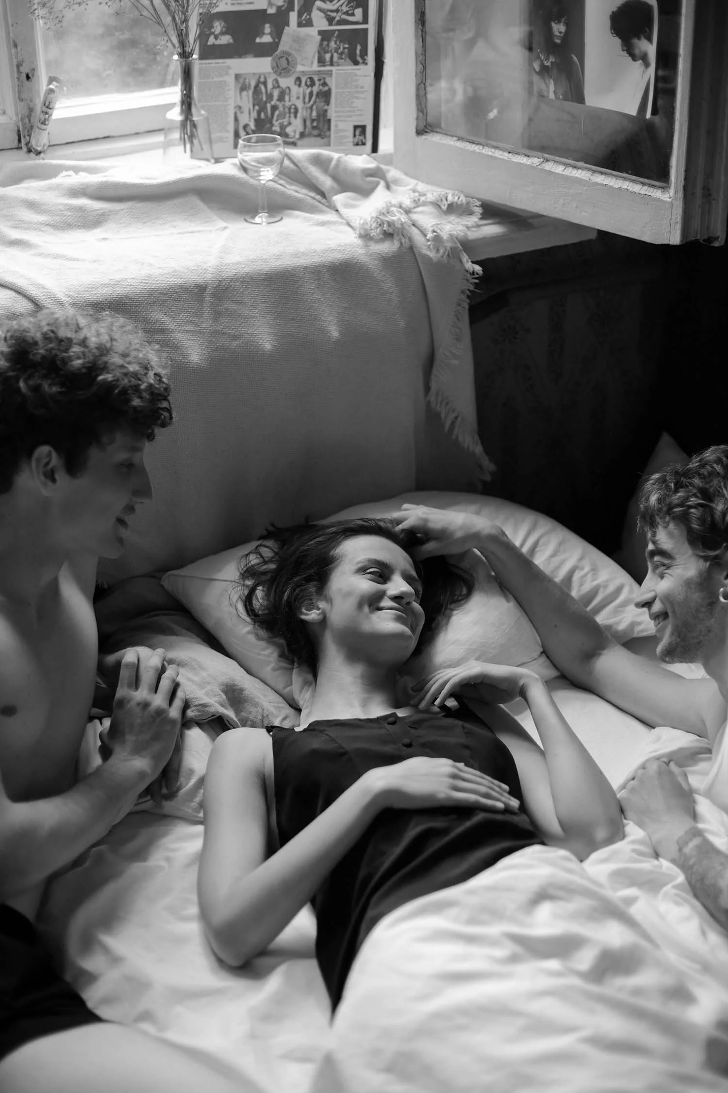 Three people are lying on a bed, smiling and interacting with each other; two men are touching the woman in the center, who is resting on a pillow, in a cozy, well-lit room.
