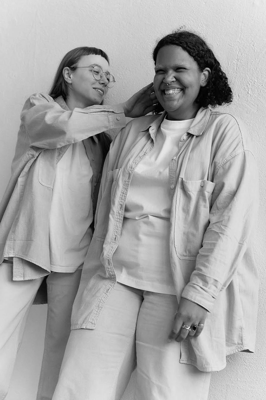 Two queer women smiling and interacting, one with glasses and short curly hair, the other with straight hair and rings on her fingers.