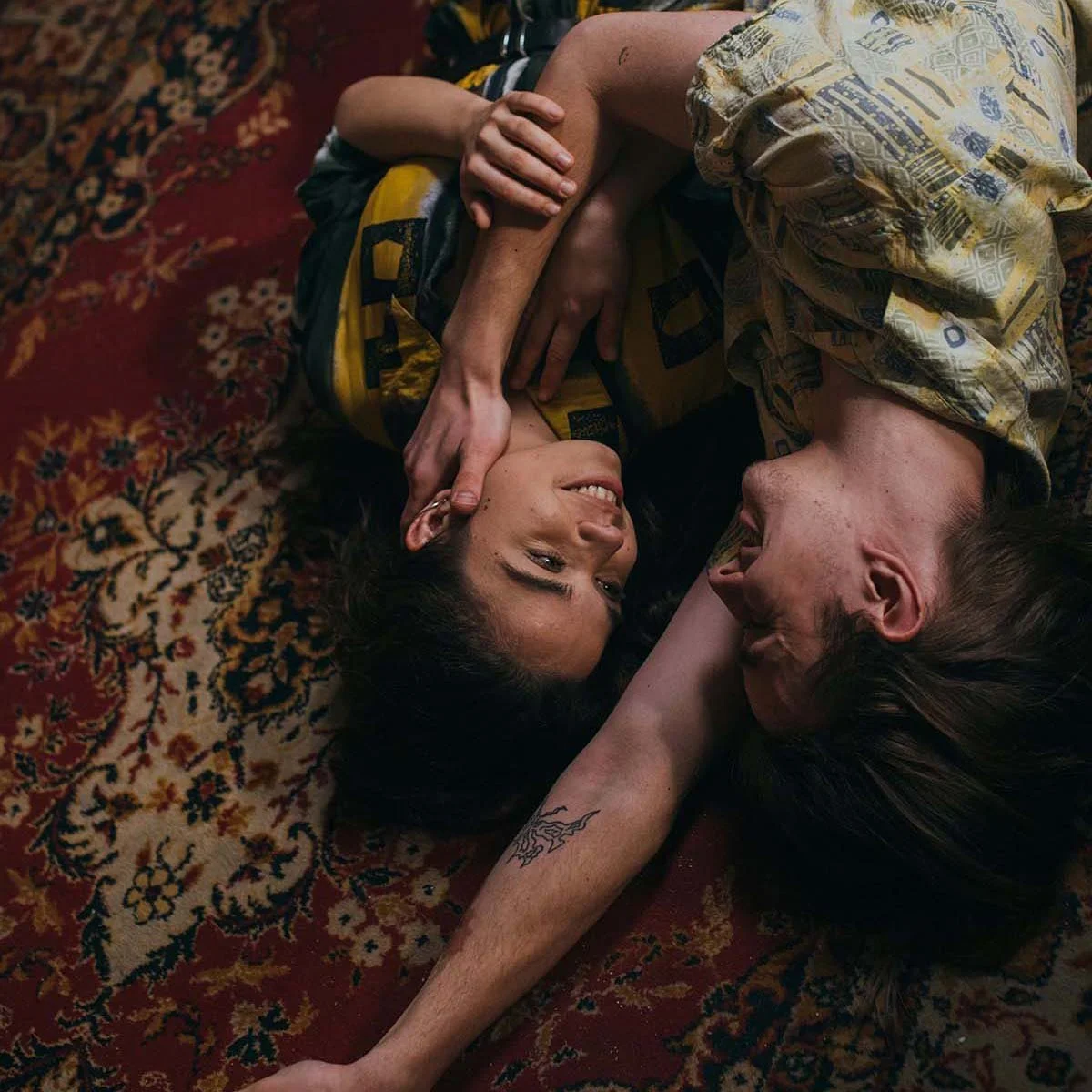 A couple lying on a patterned rug, smiling and looking at each other, with the woman’s hand gently touching the man’s face.