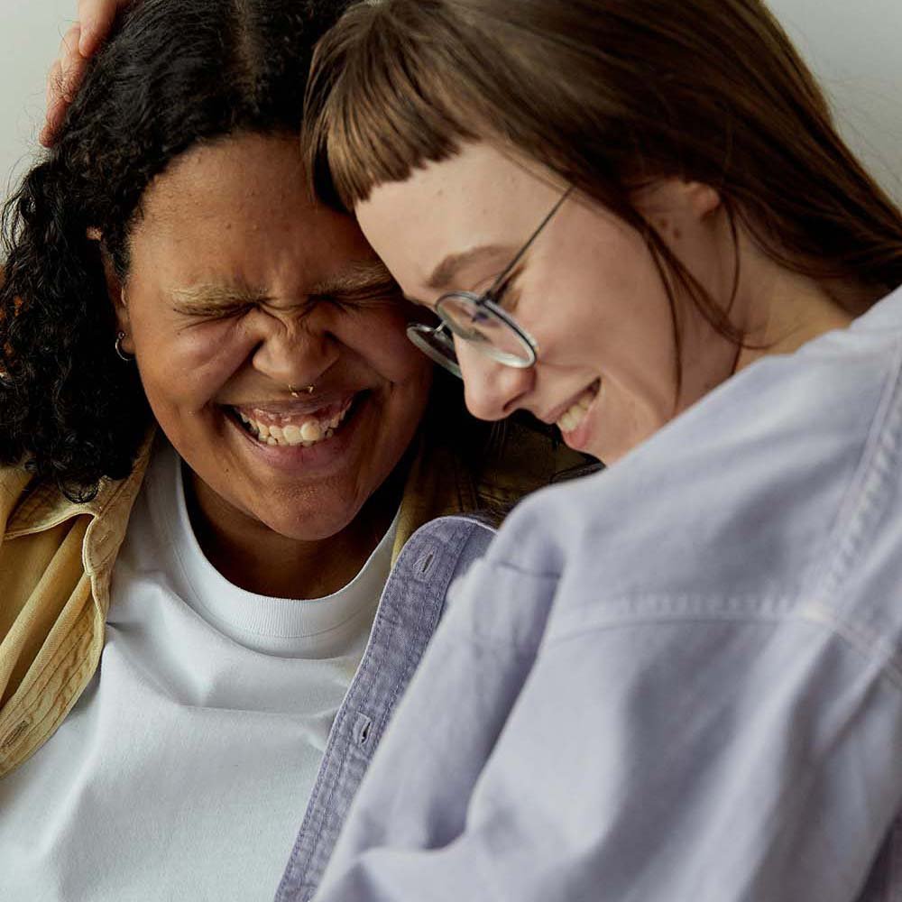 A queer couple sharing a moment of joy, their foreheads touching and smiling with eyes closed.
