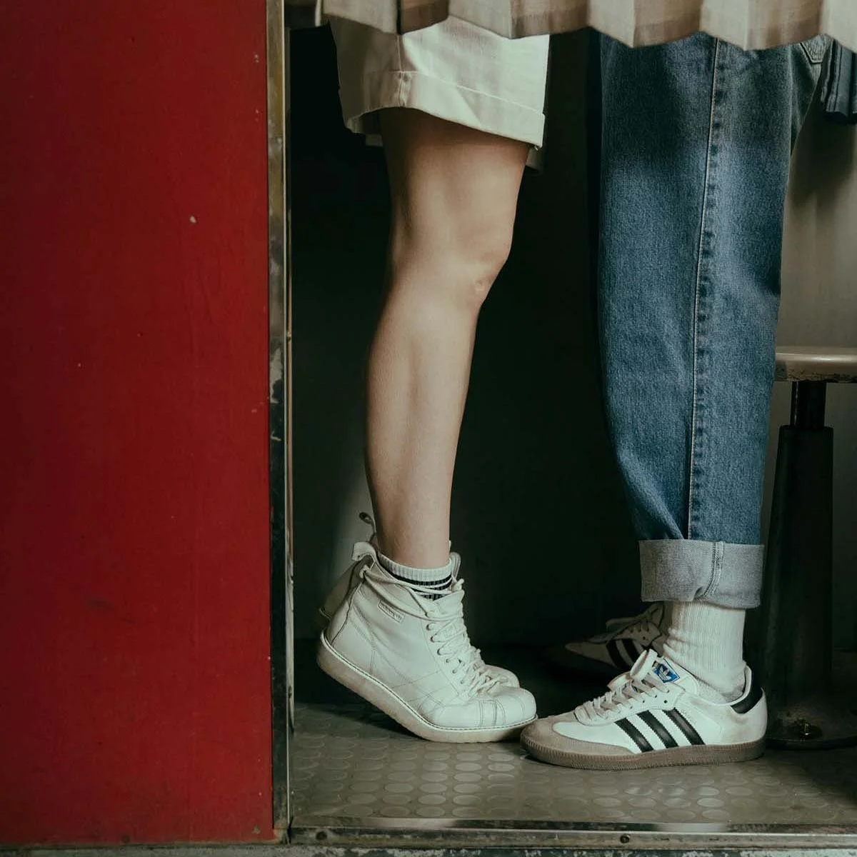 Close-up of two people standing in a photobooth with their feet touching, both wearing sneakers, one with white high-tops and the other with white athletic shoes with black stripes.