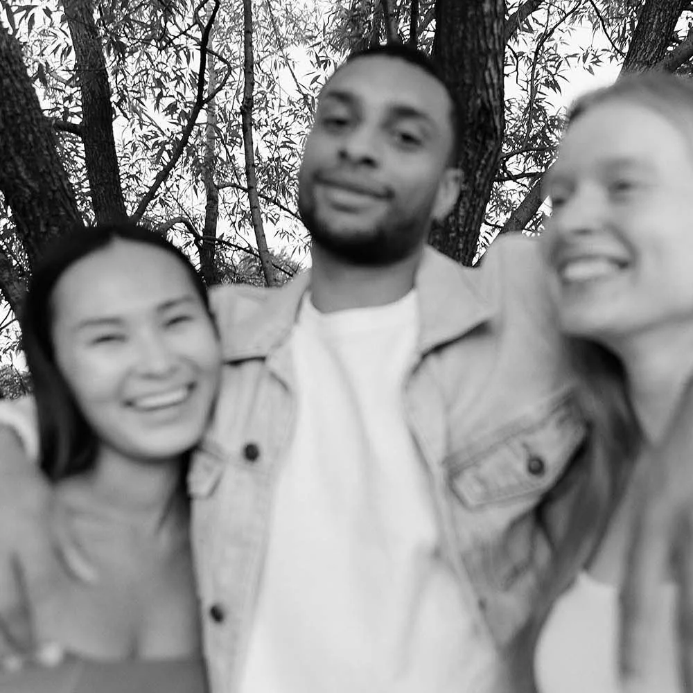 Three partners in a polyamorous relationship smiling and posing outdoors in front of trees, with two women and one man, in black-and-white photo.