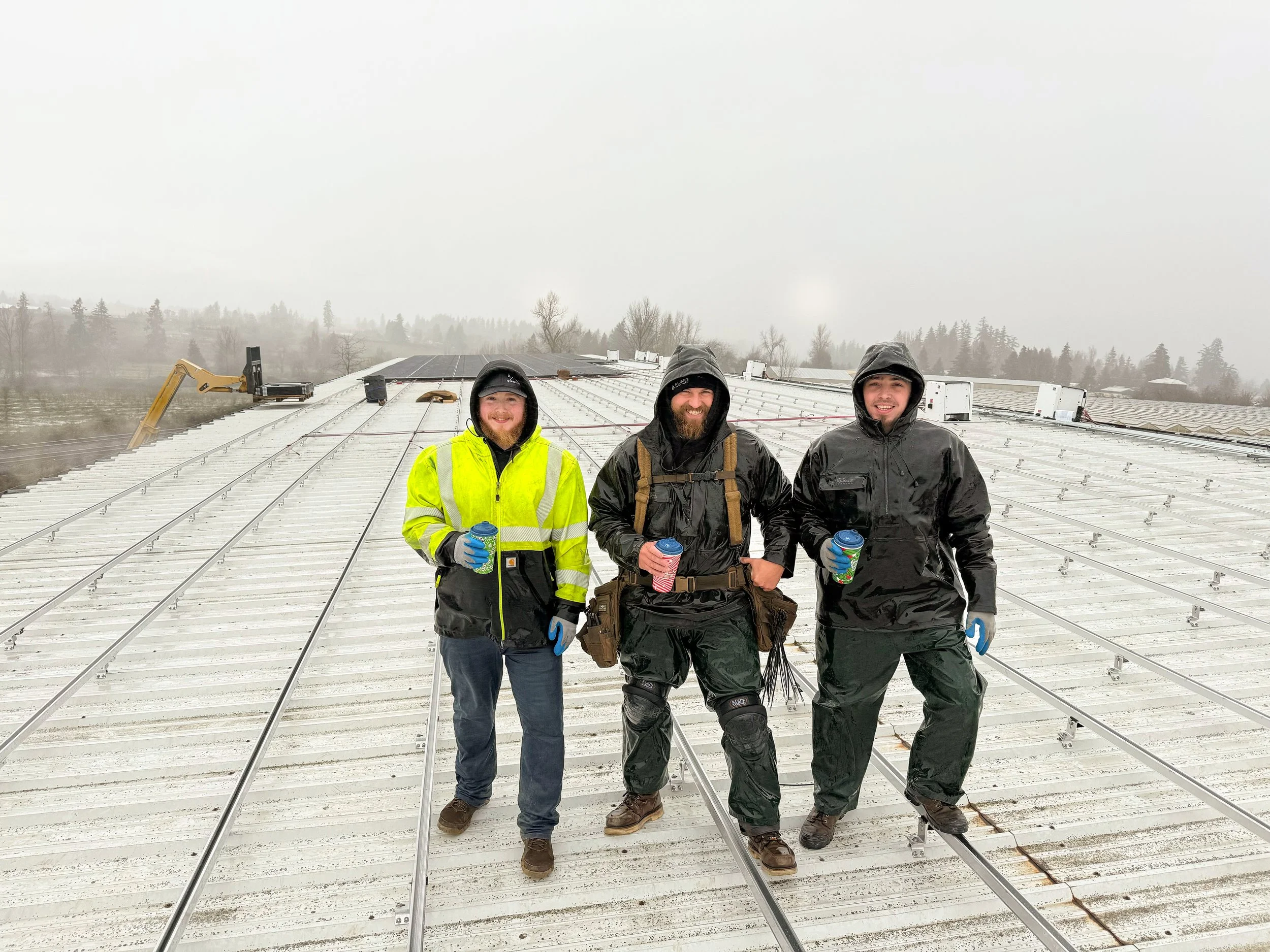 Three men standing on a metal roof in rainy weather, holding coffee cups, with a construction excavator in the background.