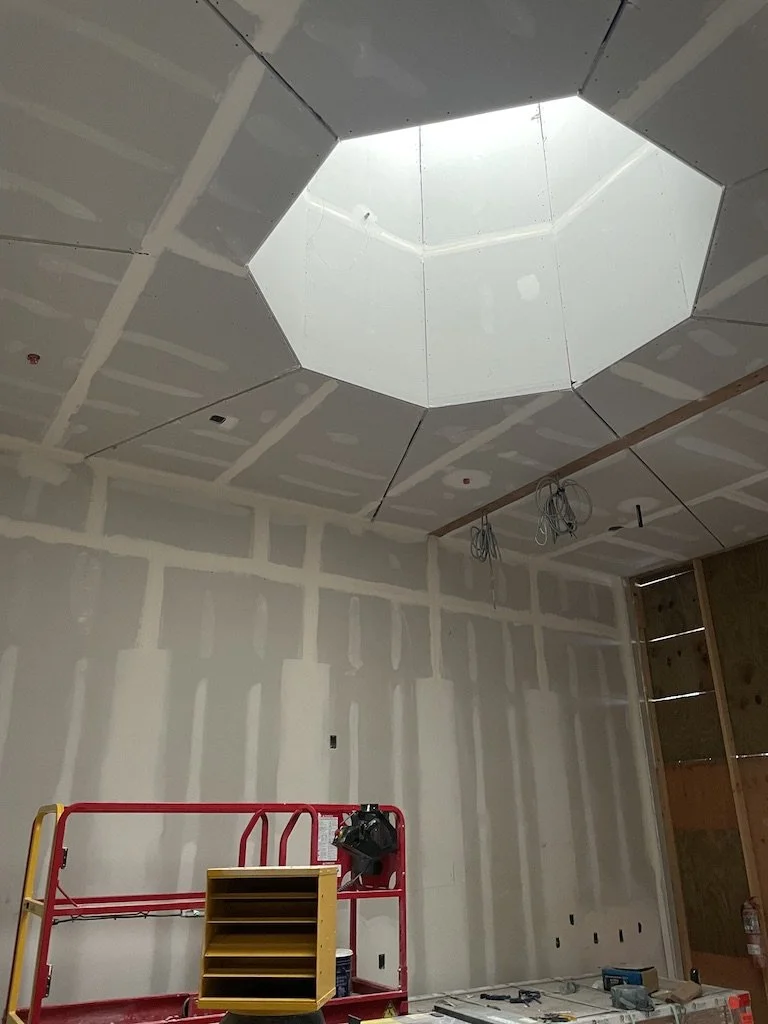 Interior of a building under construction with drywall installed, ceiling drywall has an octagonal skylight, and hanging electrical wires.