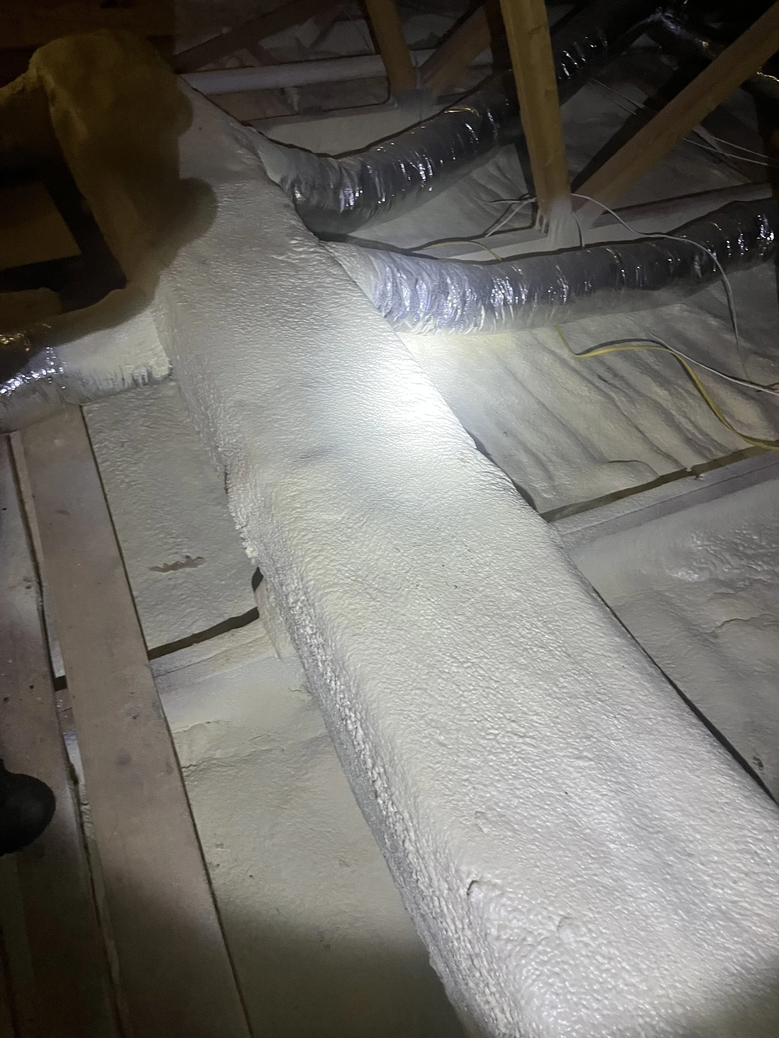 A person spray painting a wall with textured white paint in an attic space with exposed ductwork and wooden beams.