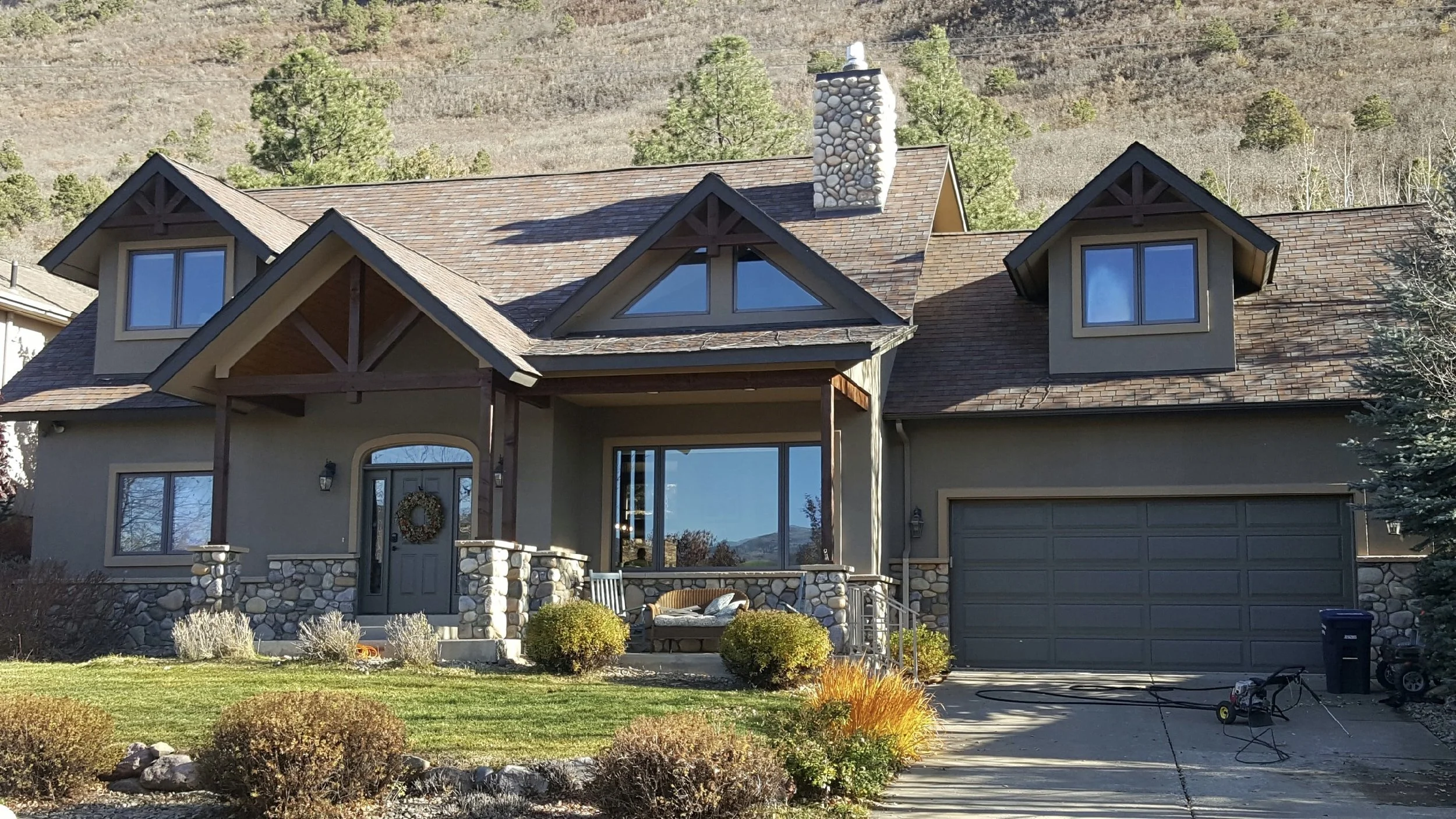 A two-story house with a stone and beige exterior, gabled roof with brown shingles, large windows, and a covered front porch decorated with a wreath. The front yard has bushes, a lawn, and garden beds, with mountains and trees in the background.