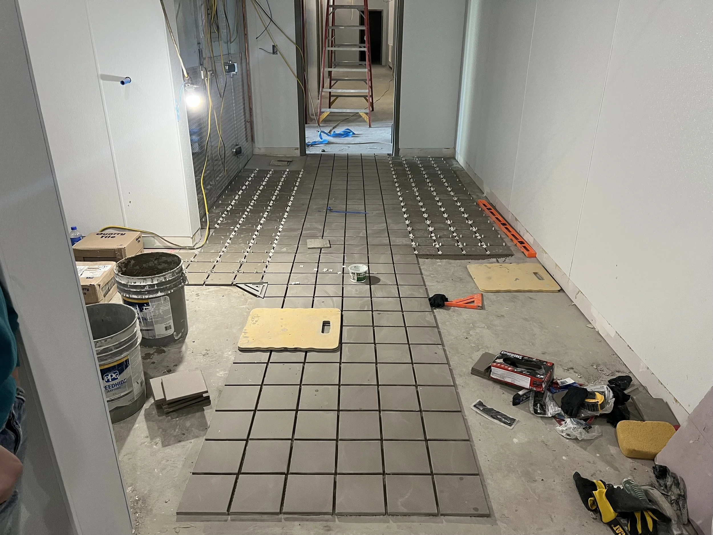 Construction site with tiles being installed on the floor, tools, buckets, and construction materials scattered around, with a ladder and wiring in the background.