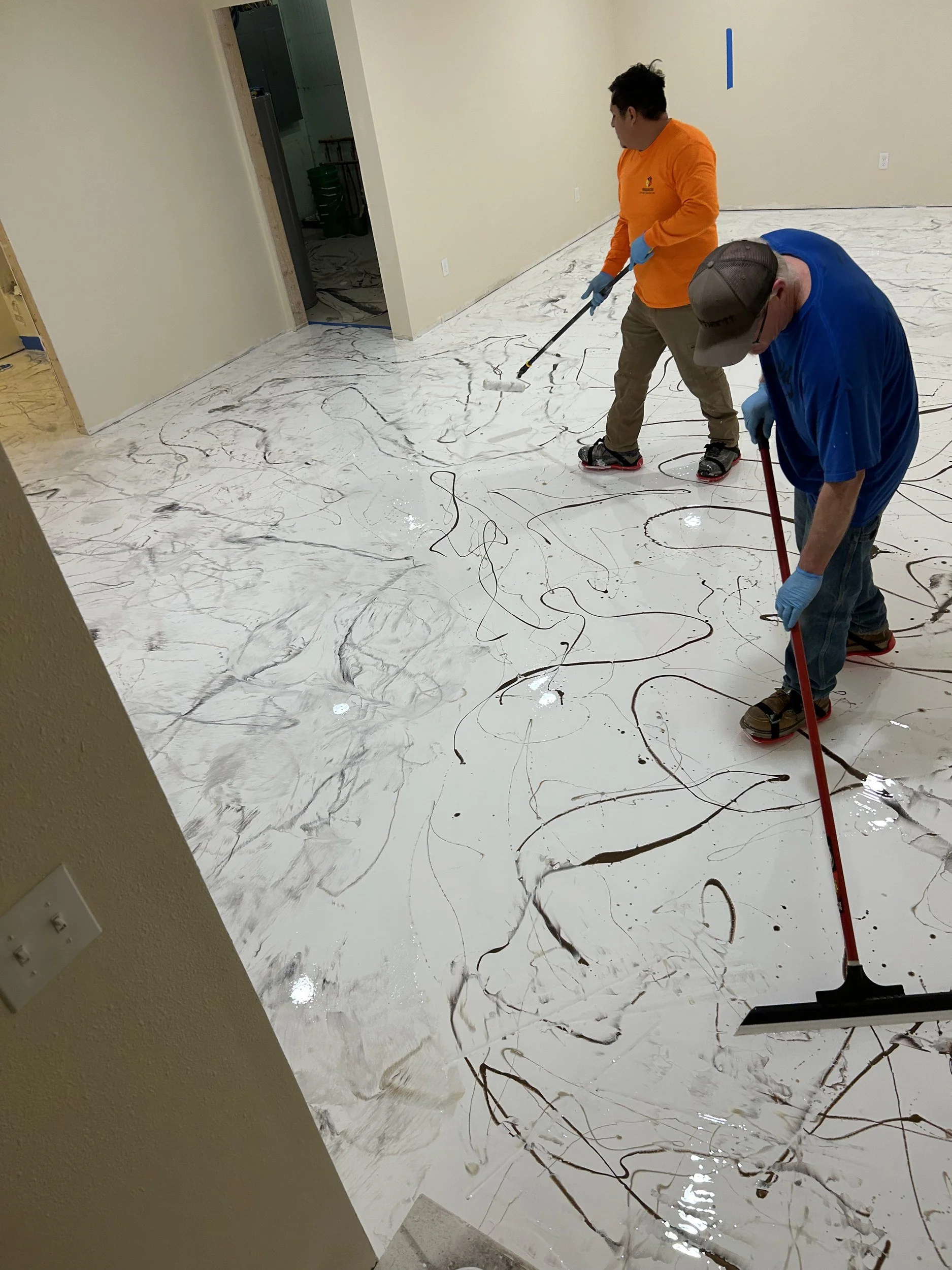 Two people are painting a floor with a white and black marble pattern, using long-handled rollers. One person is wearing an orange shirt and brown pants, and the other is wearing a blue shirt and a cap. The floor appears to be newly painted or coated.