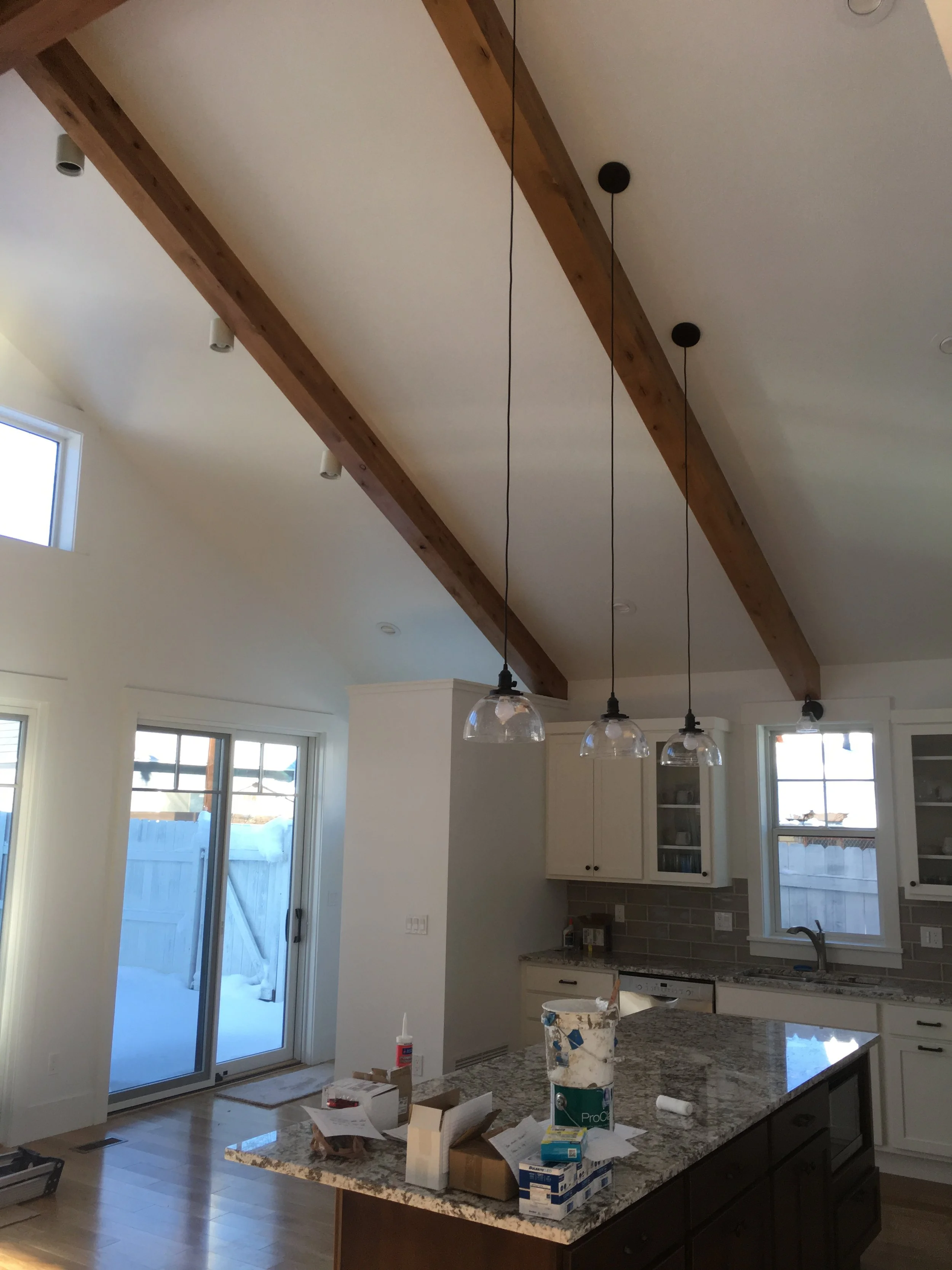 Kitchen with granite island countertop, white cabinets, and two windows. Ceiling has wooden beams with hanging lights. A sliding glass door leads outside to a snowy yard. Kitchen is under construction with tools and boxes on the island.