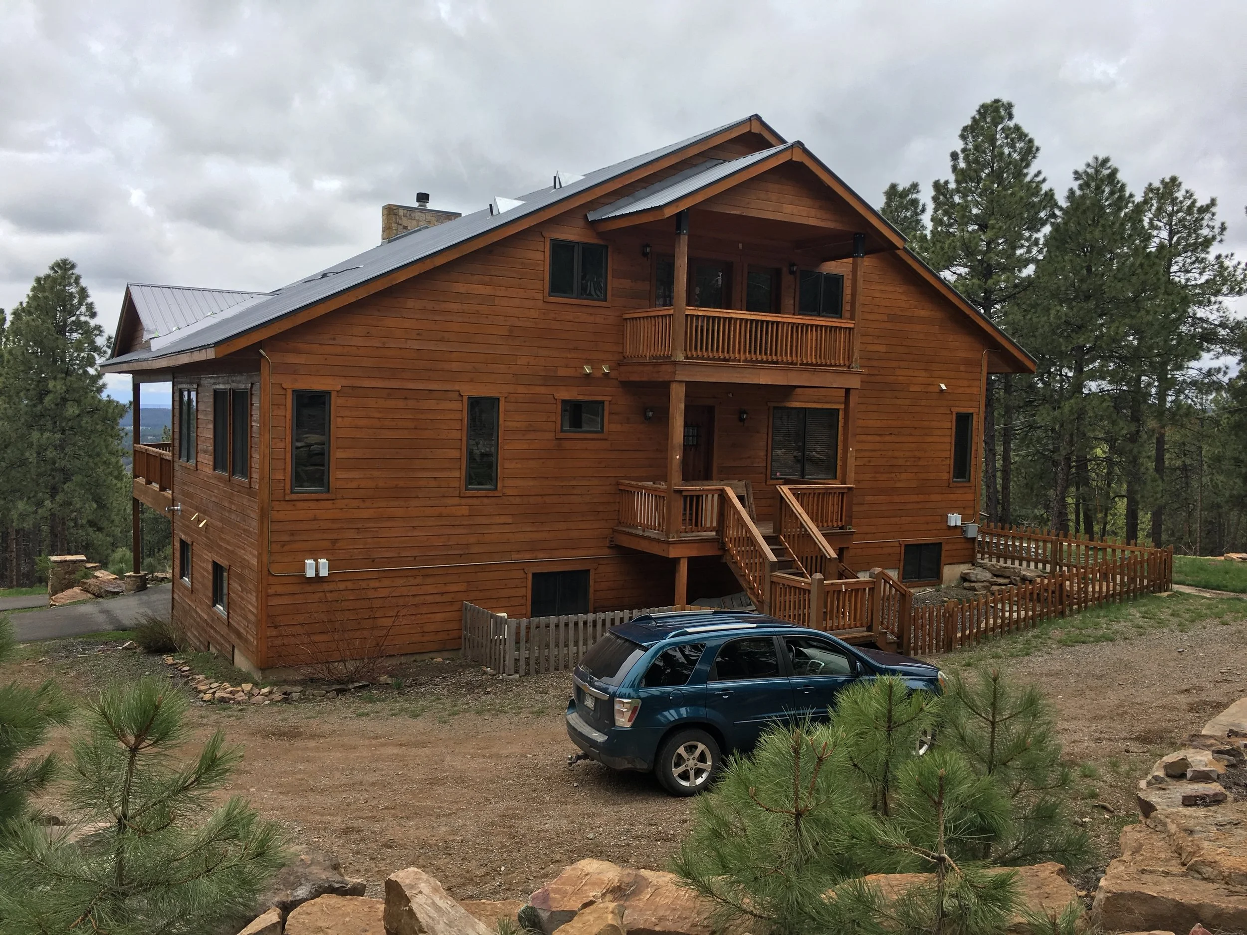A two-story wooden house with a metal roof, balconies, and stairs, set in a forested area with green trees, a gravel driveway, and a parked dark blue car.